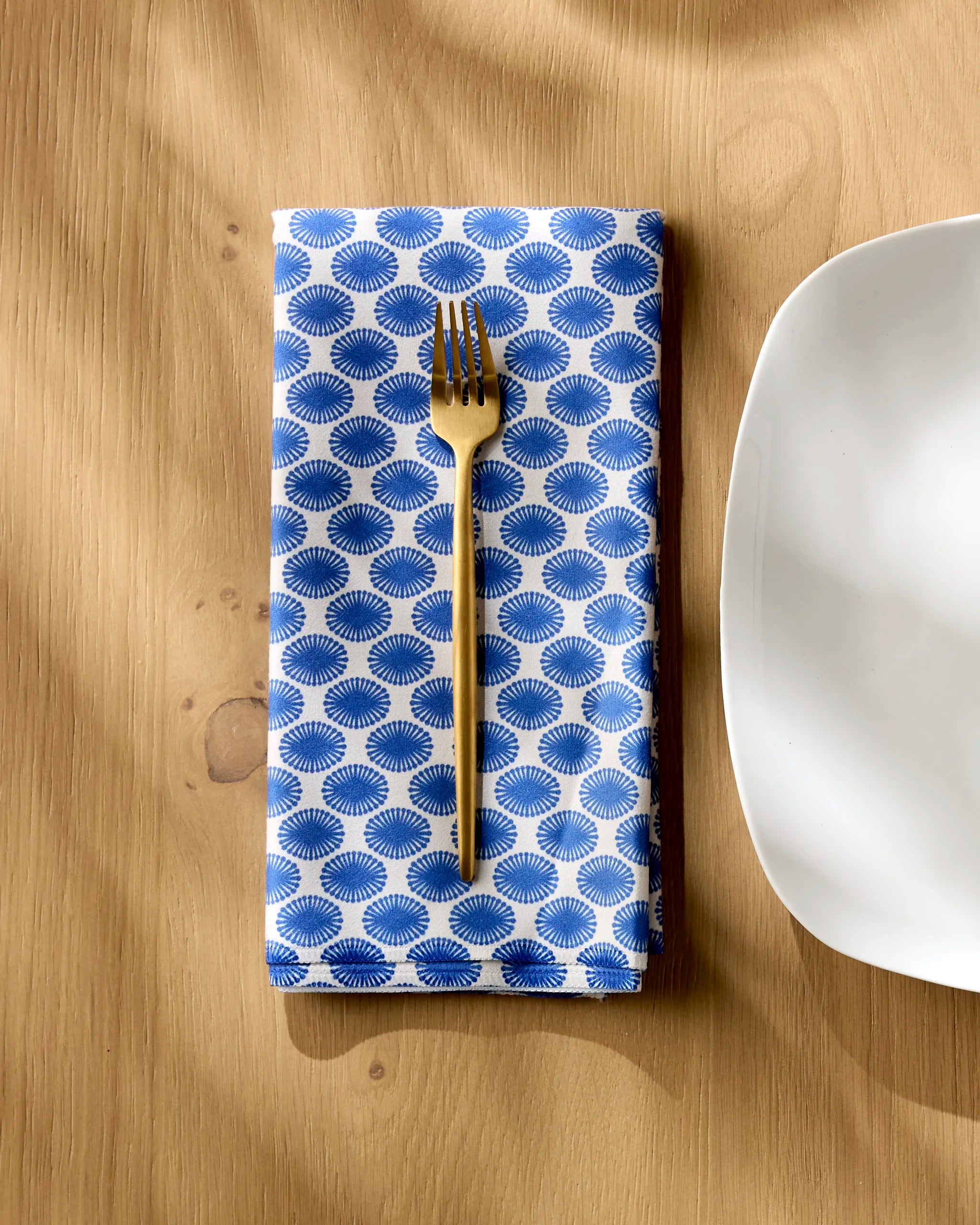 A Geometry "Lagoon" blue and white patterned reusable cloth napkin sits beside a gold fork and white square plate on a light wooden table, sunlight casting soft shadows over the scene.