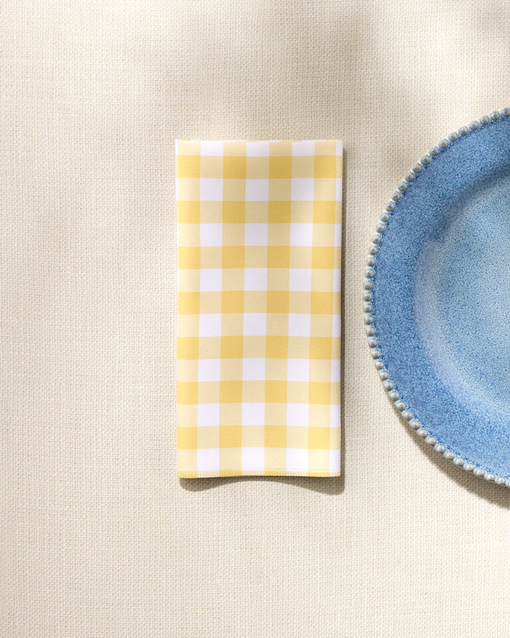 The GEOMETRY Lemon Gingham washable dinner napkin, in yellow and white checks, is neatly folded beside a blue plate with a textured rim on a light beige tablecloth.