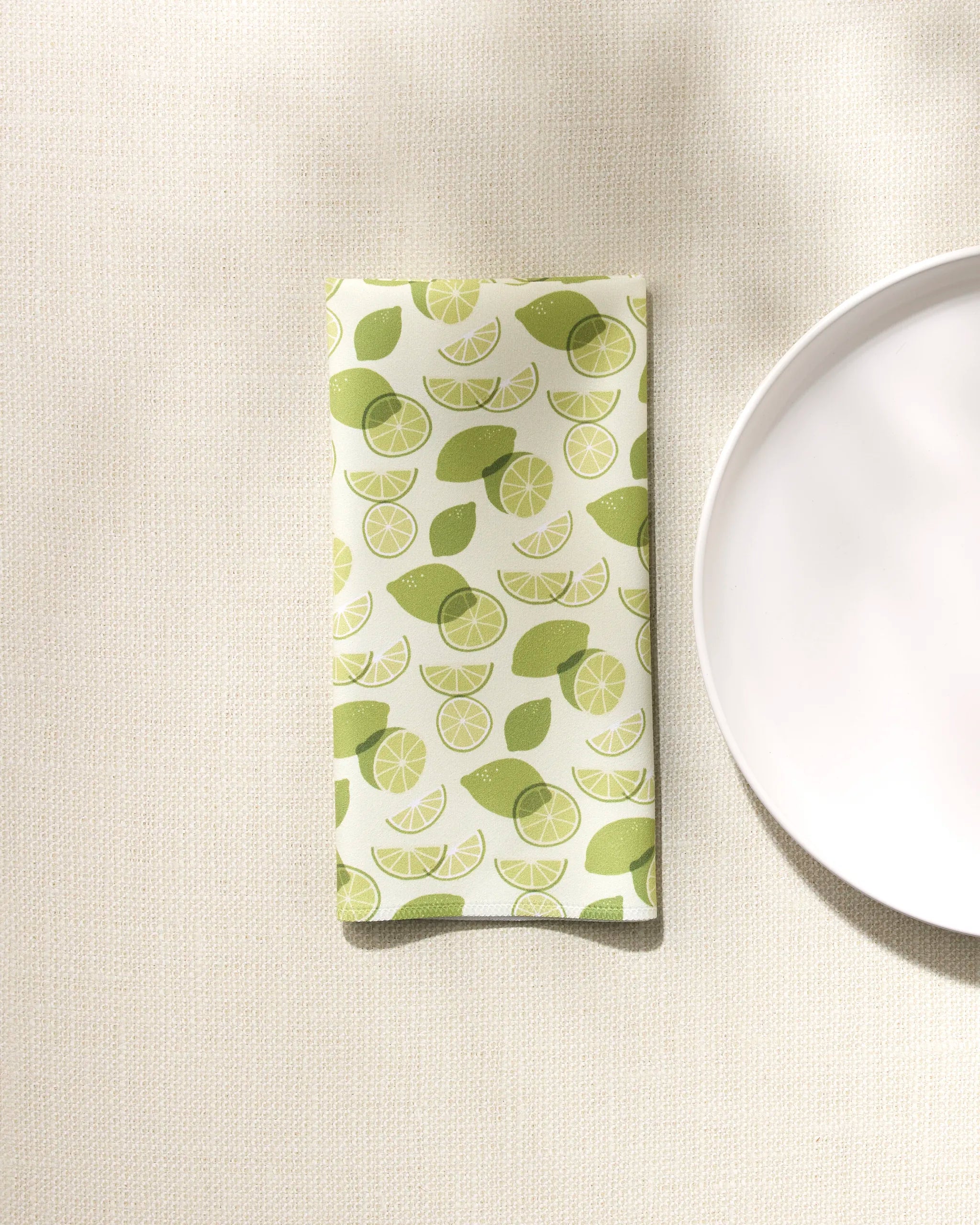 A folded GEOMETRY "Limes" eco-friendly napkin with a green lime fruit and slice design rests on a textured beige surface beside a white plate.