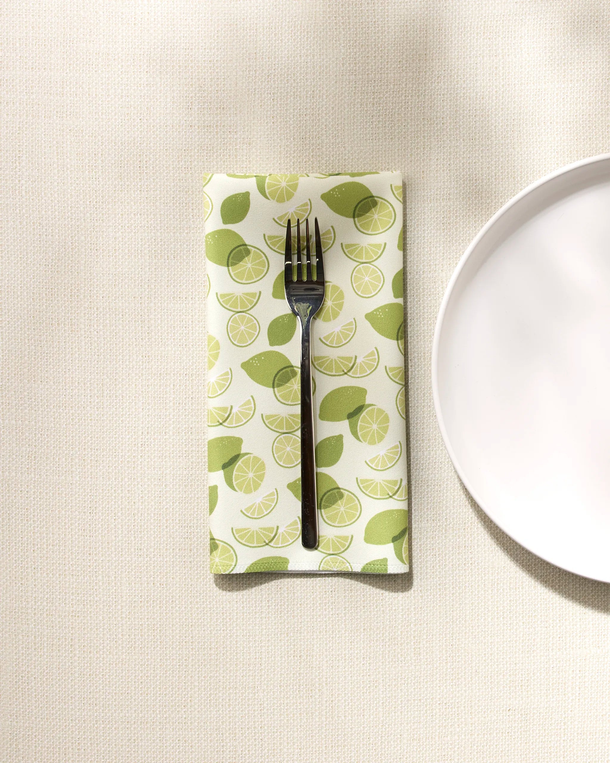 A silver fork rests on a GEOMETRY eco-friendly napkin featuring a limes pattern, set on a textured tablecloth beside a white plate partially visible to the right.