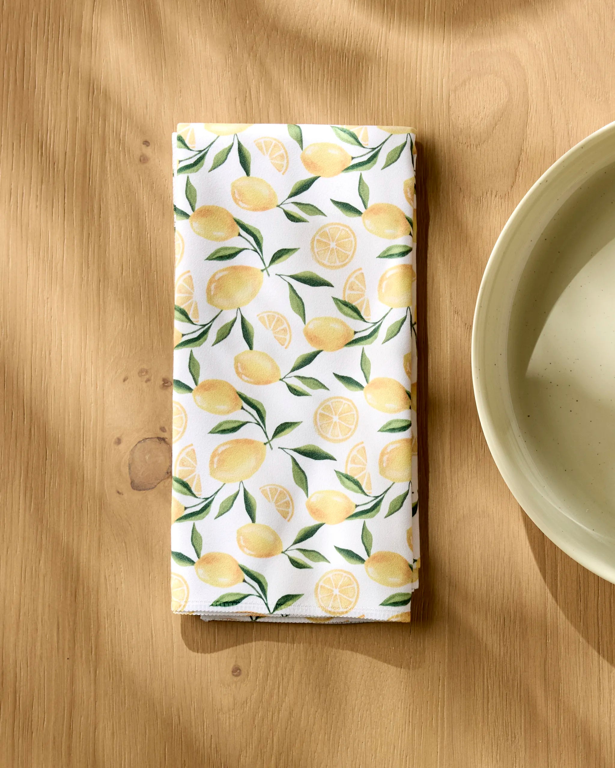 A Geometry Limoncello white cloth napkin, featuring yellow lemons and green leaves, is neatly folded on a light wooden table beside a cream ceramic bowl. Sunlight casts gentle shadows over the scene.