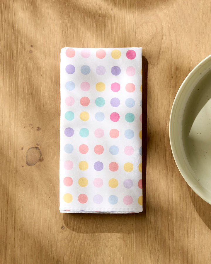 The "Lots of Dots" eco-friendly napkin by Rebecca Bobko, featuring pastel polka dots on white, rests on a light wooden table beside a cream plate.