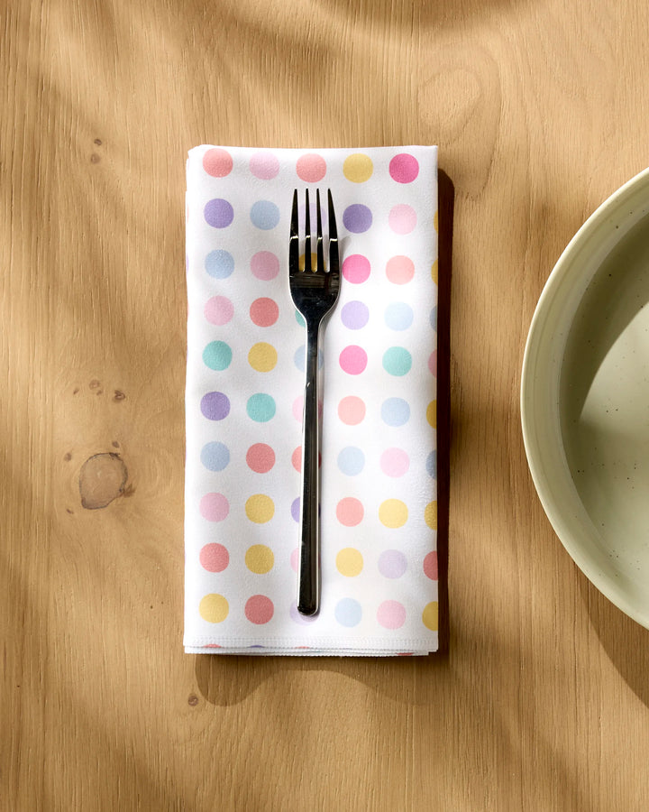 A silver fork rests on a folded Rebecca Bobko Lots of Dots napkin, an eco-friendly dinner napkin decorated with pastel polka dots, set on a light wooden table beside a beige plate.