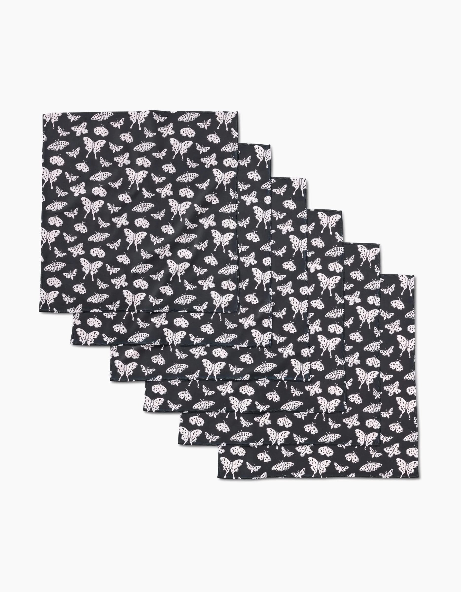 Five folded Kayla Ann Lunar Moths Black dinner napkins, crafted from Geosuede with a repeating white moth pattern, are displayed in a staggered stack on a plain white background.