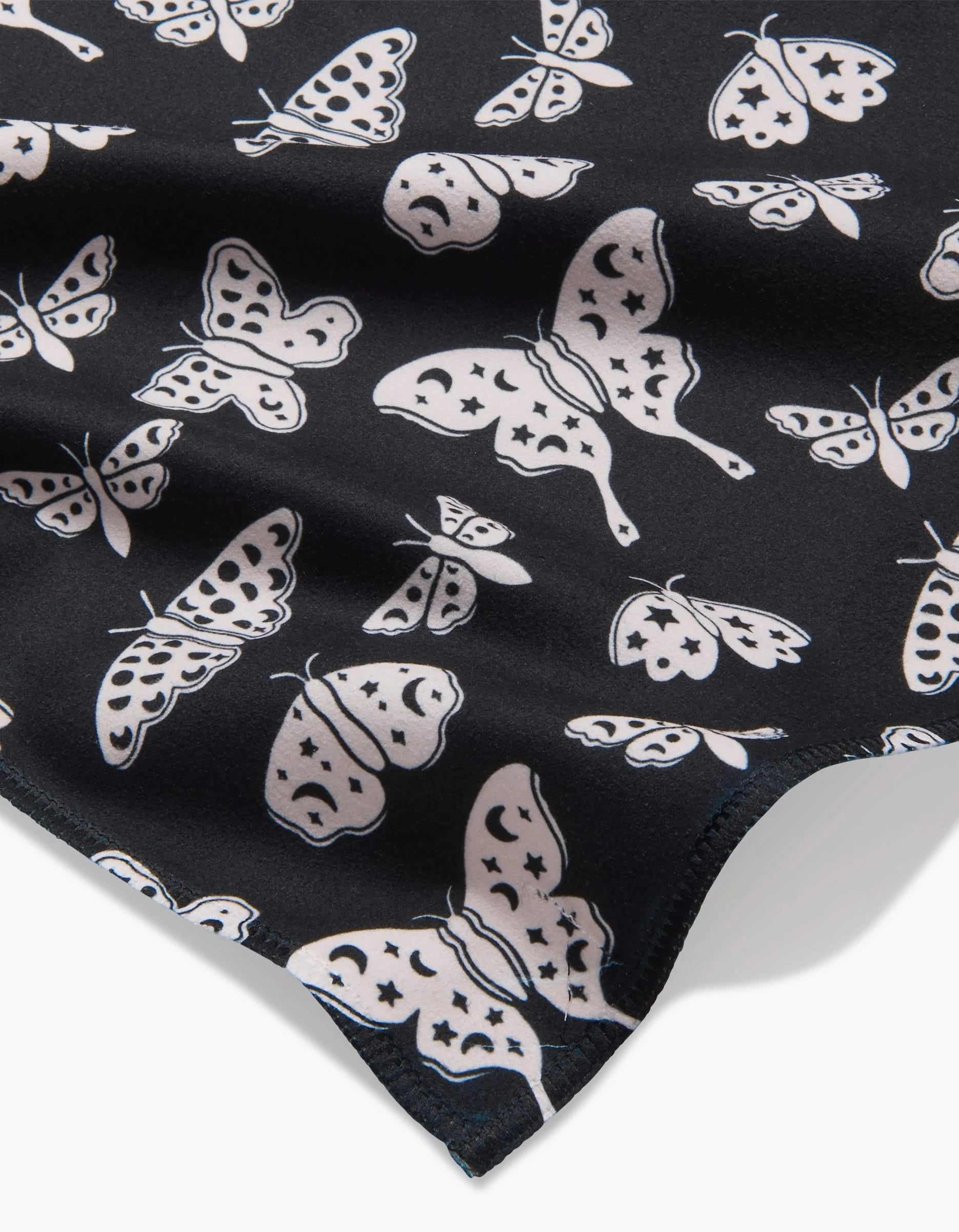 A close-up of Kayla Anns Lunar Moths Black shows black fabric with white butterfly and moth illustrations in various sizes and styles, with the edge of the fabric slightly folded.