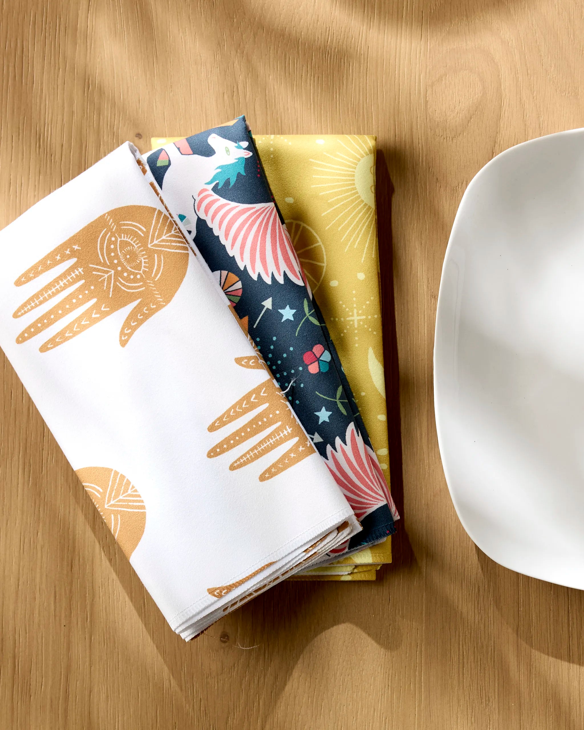 Three folded Magical Moments reusable cloth napkins by Jessica Swift—featuring white with brown hands, dark with stars and wings, and yellow with suns—are arranged next to a white plate on a light wooden table.