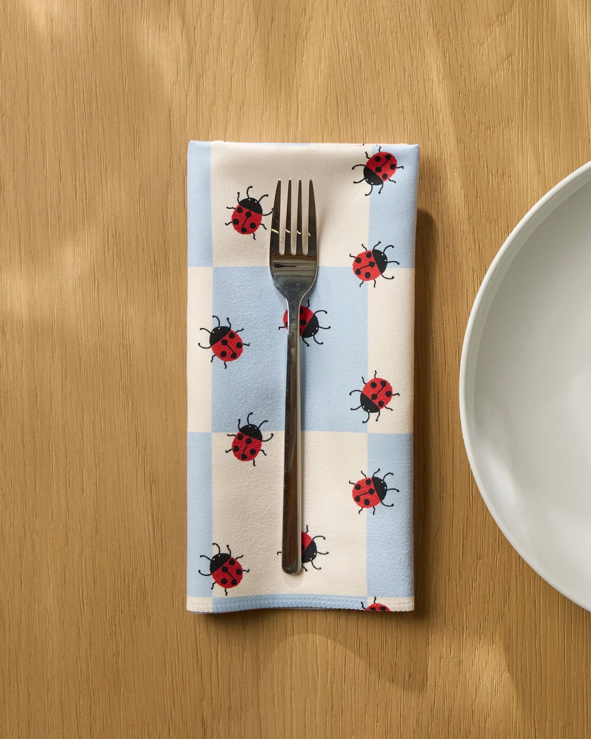 A fork rests on Geometry's Mariquita Pop, a blue and white checkered reusable napkin with red ladybugs, placed on a wooden table next to a white plate.