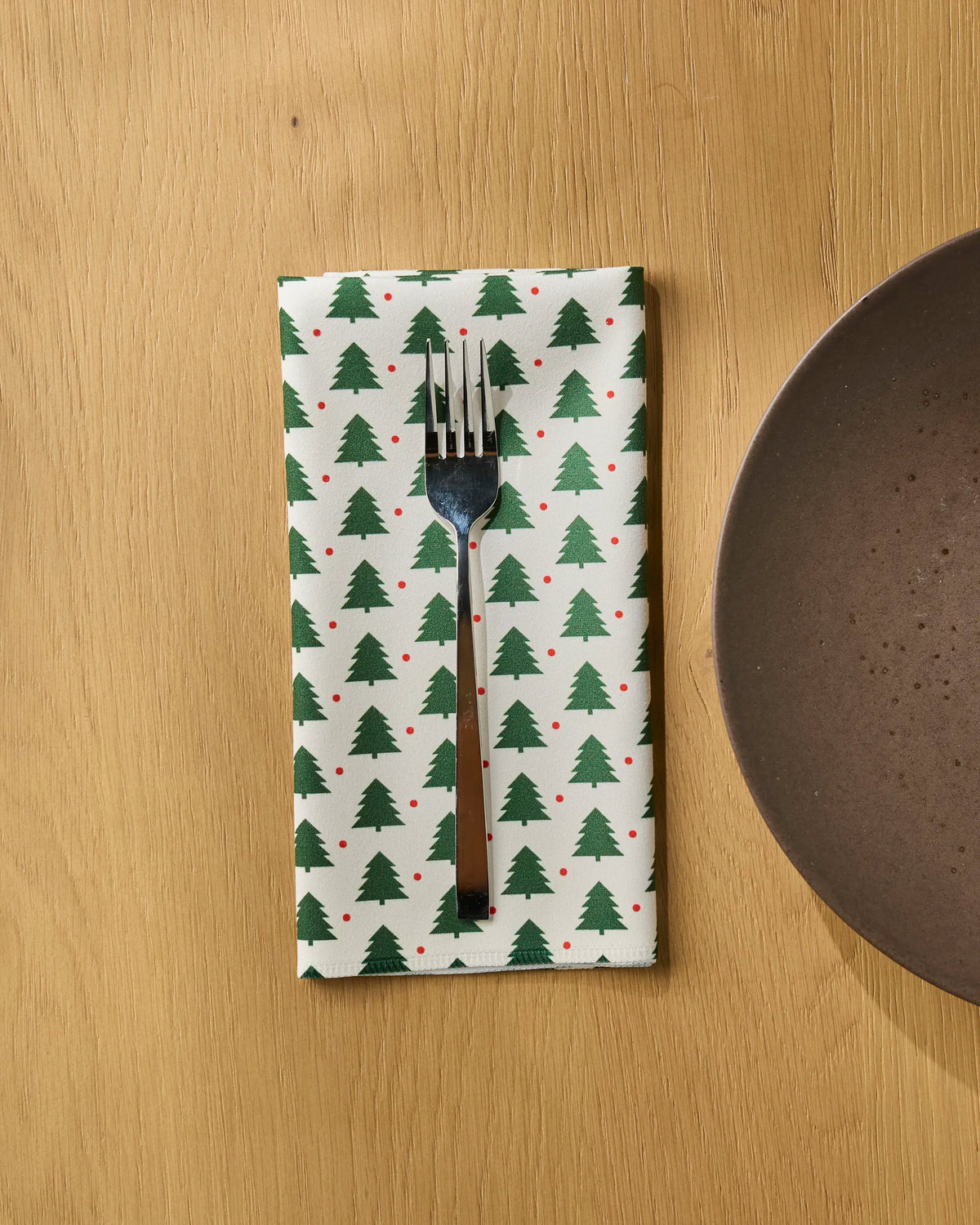 A fork rests on a Geometry Merry Noel recycled dinner napkin, which features green Christmas trees and red dots, placed on a light wooden table next to a brown plate.