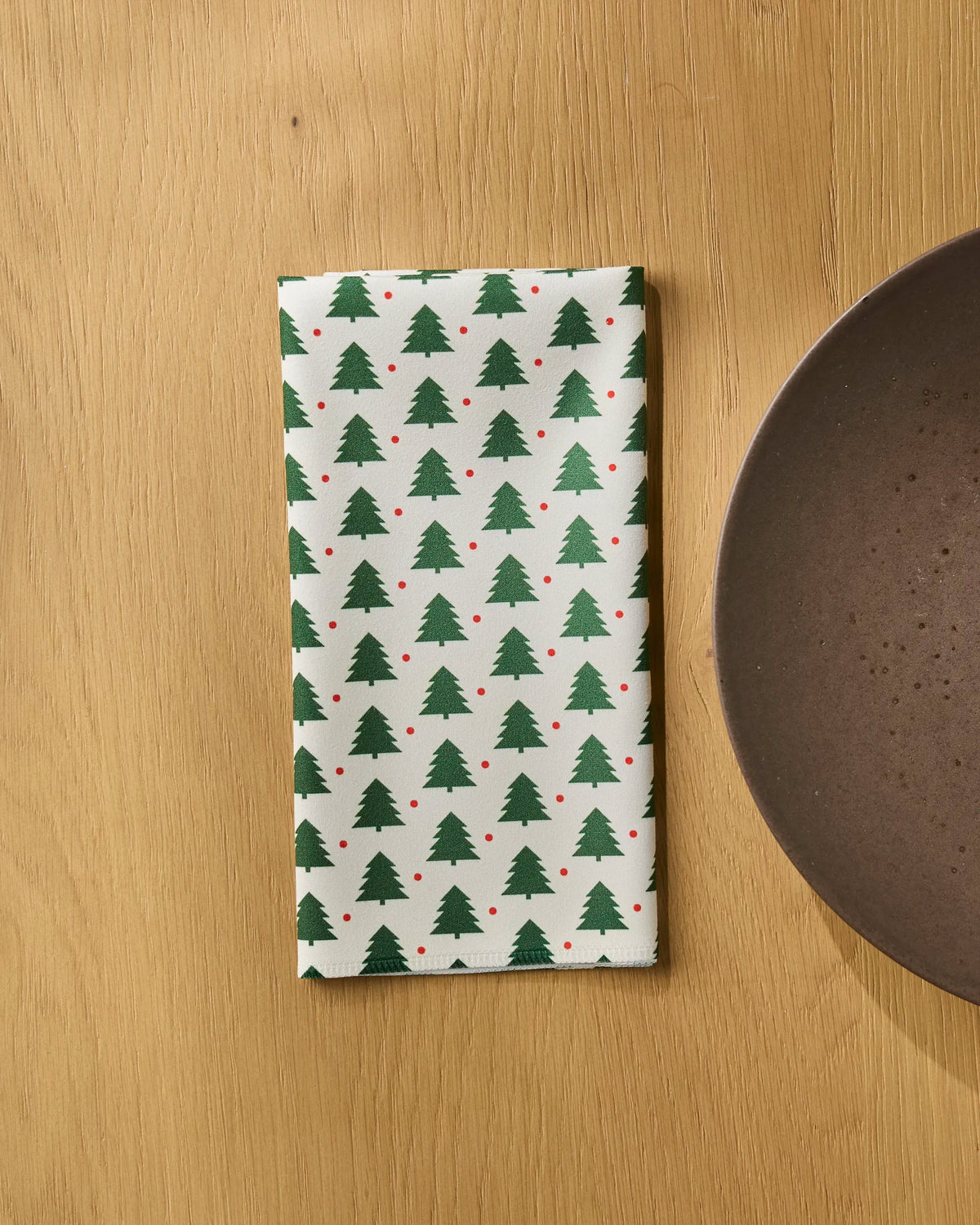 A folded napkin from the Merry Noel Dinner Napkin Set by Geometry, adorned with green Christmas tree patterns and small red dots, rests on a wooden table beside a brown ceramic plate. These absorbent cloth napkins bring festive charm to your holiday table setting.