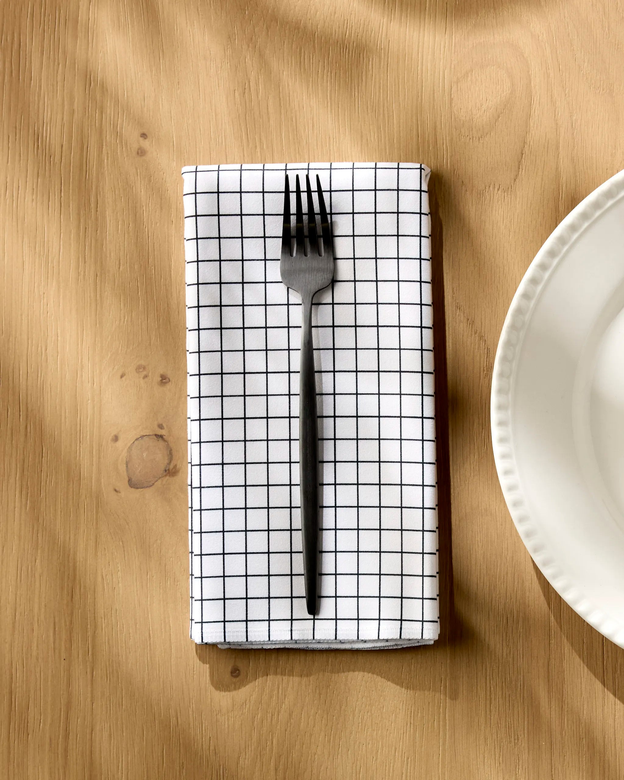 A metal fork sits on the Geometry Metro Grid reusable cloth napkin, featuring a black grid pattern, atop a light wooden table beside a white scalloped plate.