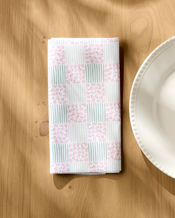 A Hufton Studio Mini Flowers and Stripes Check folded cloth napkin, featuring a pink floral and light blue striped checkerboard pattern, sits on a wooden table beside a scalloped white plate.