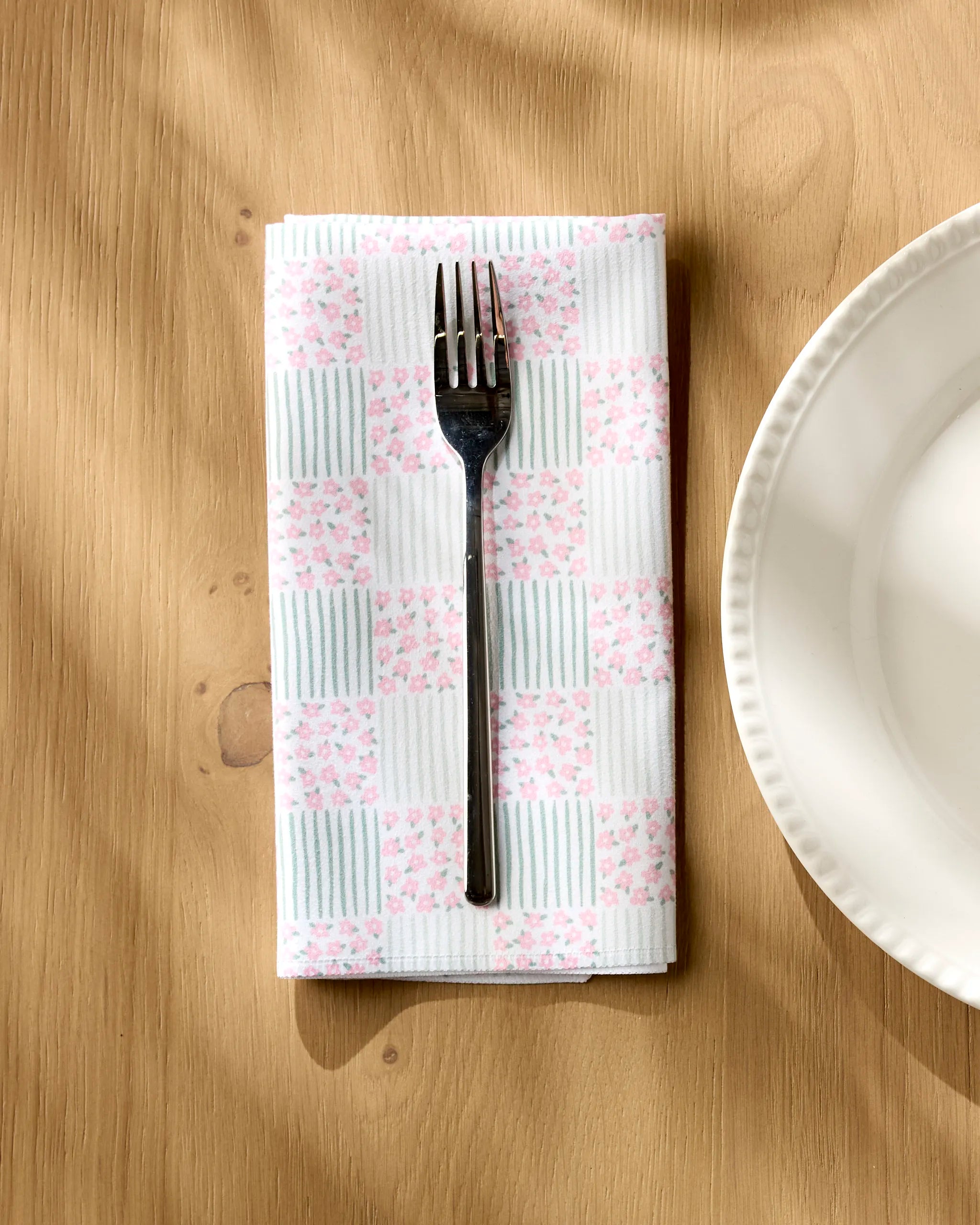 A metal fork sits on a folded Hufton Studio Mini Flowers and Stripes Check napkin beside a white scalloped plate on a light wood table.