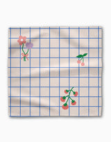 The Mini Harvest Dinner Napkin Set by Letters By Shells features beige fabric with a blue grid pattern, simple pink and purple flower illustrations, cherries, and tomato vines. Slightly wrinkled, this sustainable table linen adds charm to any setting.