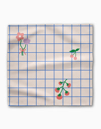 The Mini Harvest Dinner Napkin Set by Letters By Shells features beige fabric with a blue grid pattern, simple pink and purple flower illustrations, cherries, and tomato vines. Slightly wrinkled, this sustainable table linen adds charm to any setting.