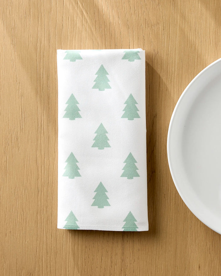 A Kailee Wright "Mini Trees" white Geosuede fabric dinner napkin featuring green pine trees is neatly folded on a wooden table beside a white plate.