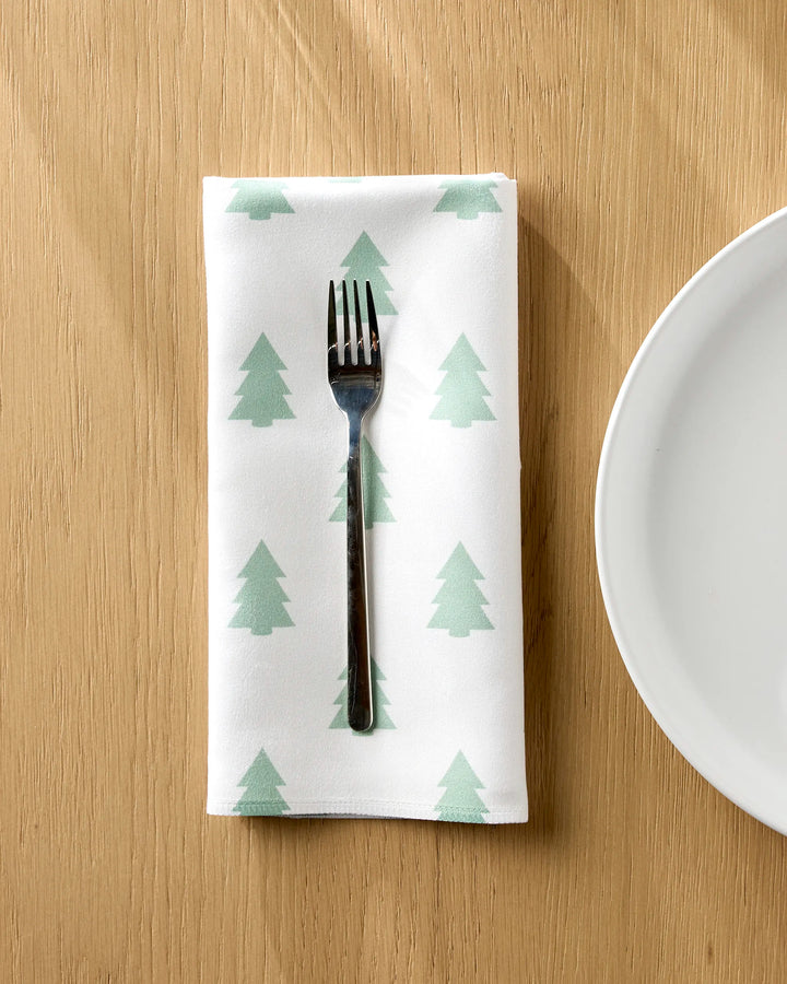 A metal fork rests on a Kailee Wright Mini Trees white Geosuede napkin with light green tree patterns, set on a wooden table next to a white plate.