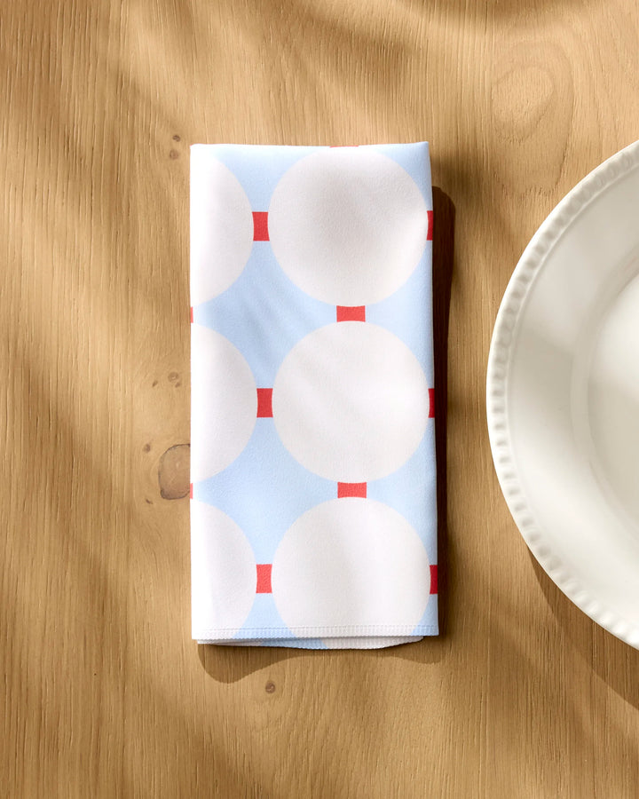 The Mist Blue Oasis reusable dinner napkin by Happy Hemera, featuring a light blue and white geometric pattern with red squares, is placed on a wooden table next to a white ceramic plate.