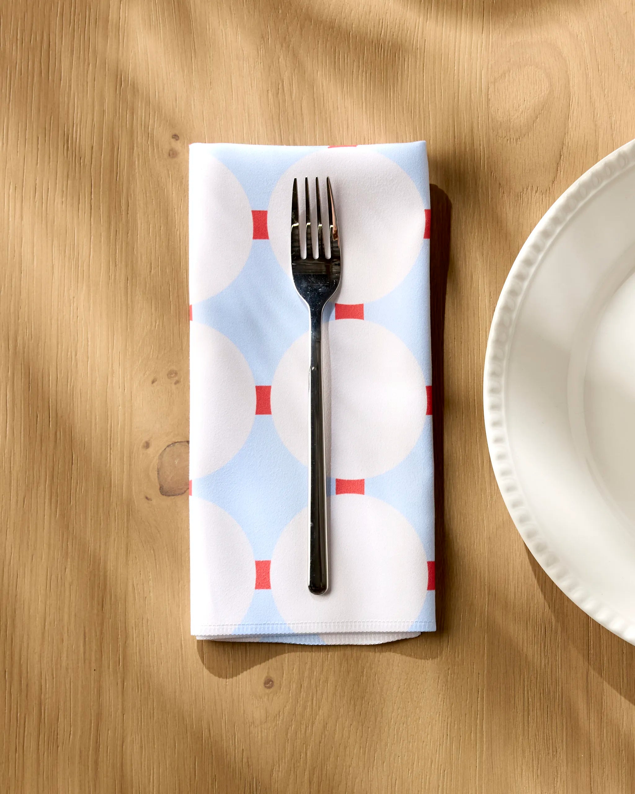 A silver fork rests on Happy Hemera’s Mist Blue Oasis dinner napkin, adorned with white circles and red lines, placed on a wooden table beside a scalloped white plate.