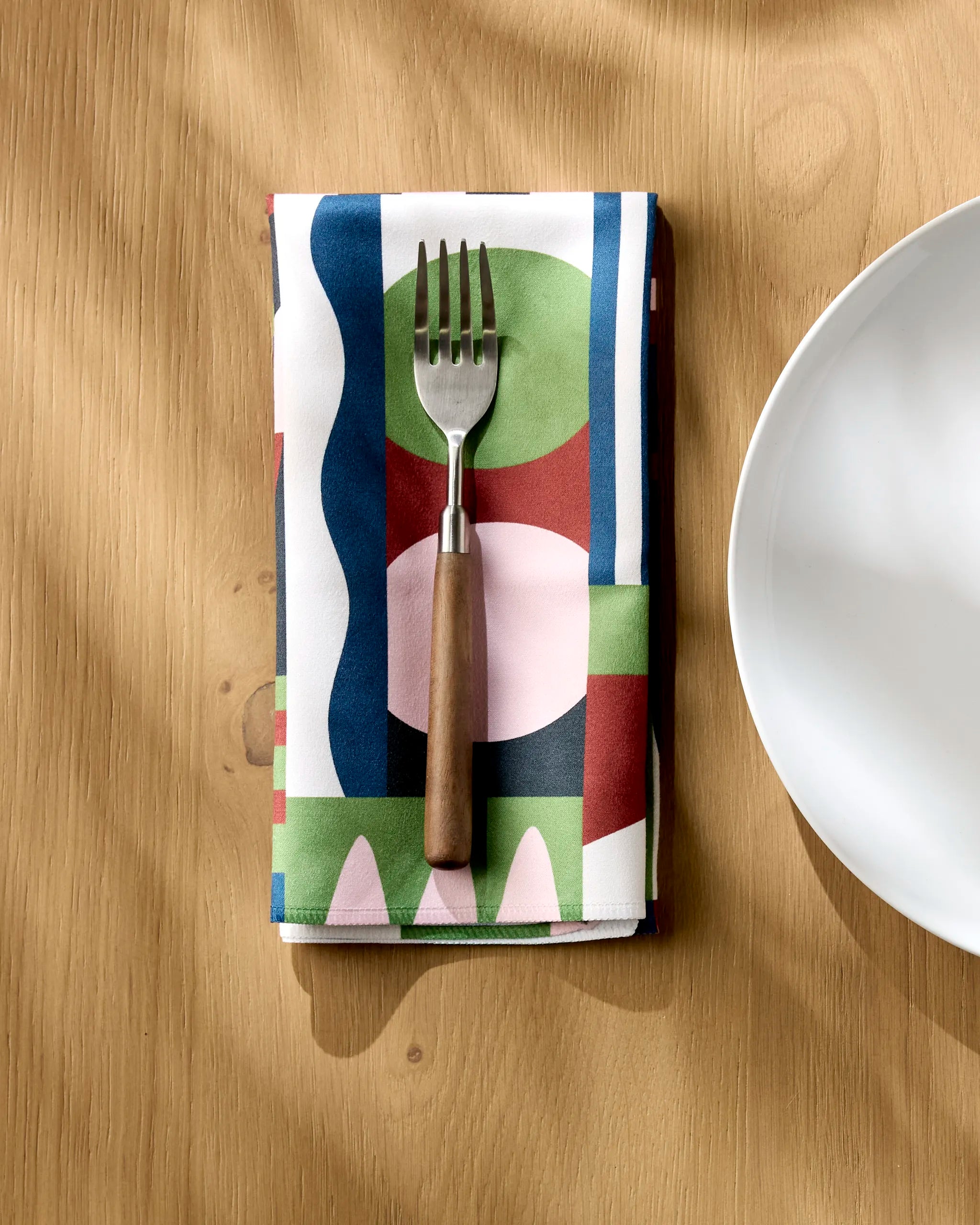 A fork with a wooden handle rests on a Claire Iglesias Modern Bauhaus Mood napkin, its colorful geometric pattern bright beside a white plate on a light wood table as sunlight casts soft shadows across the scene.