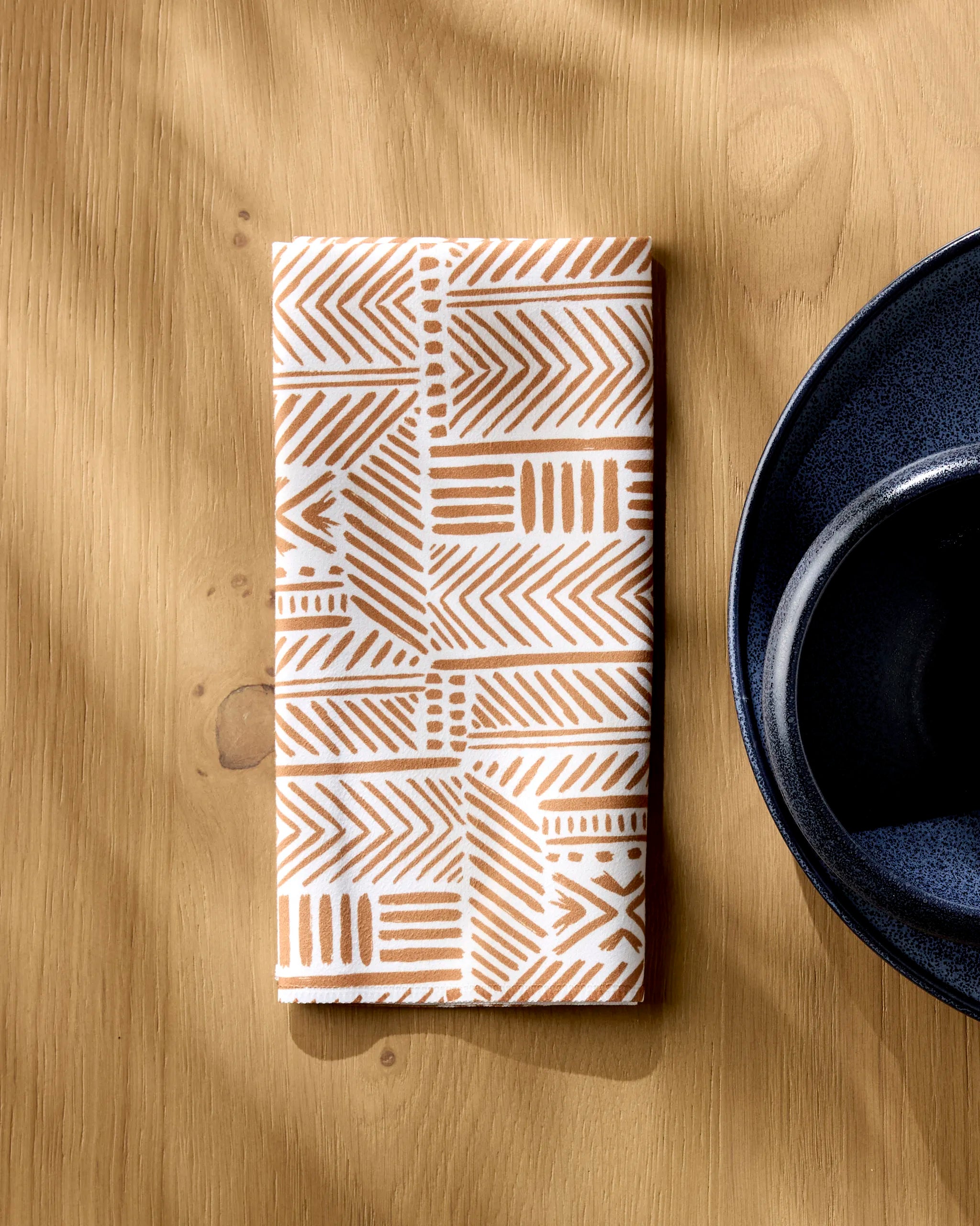 A folded Mud Cloth Natural napkin by Hufton Studio, featuring a brown geometric pattern, rests on a light wood table beside a dark blue bowl as sunlight casts soft shadows across the surface.