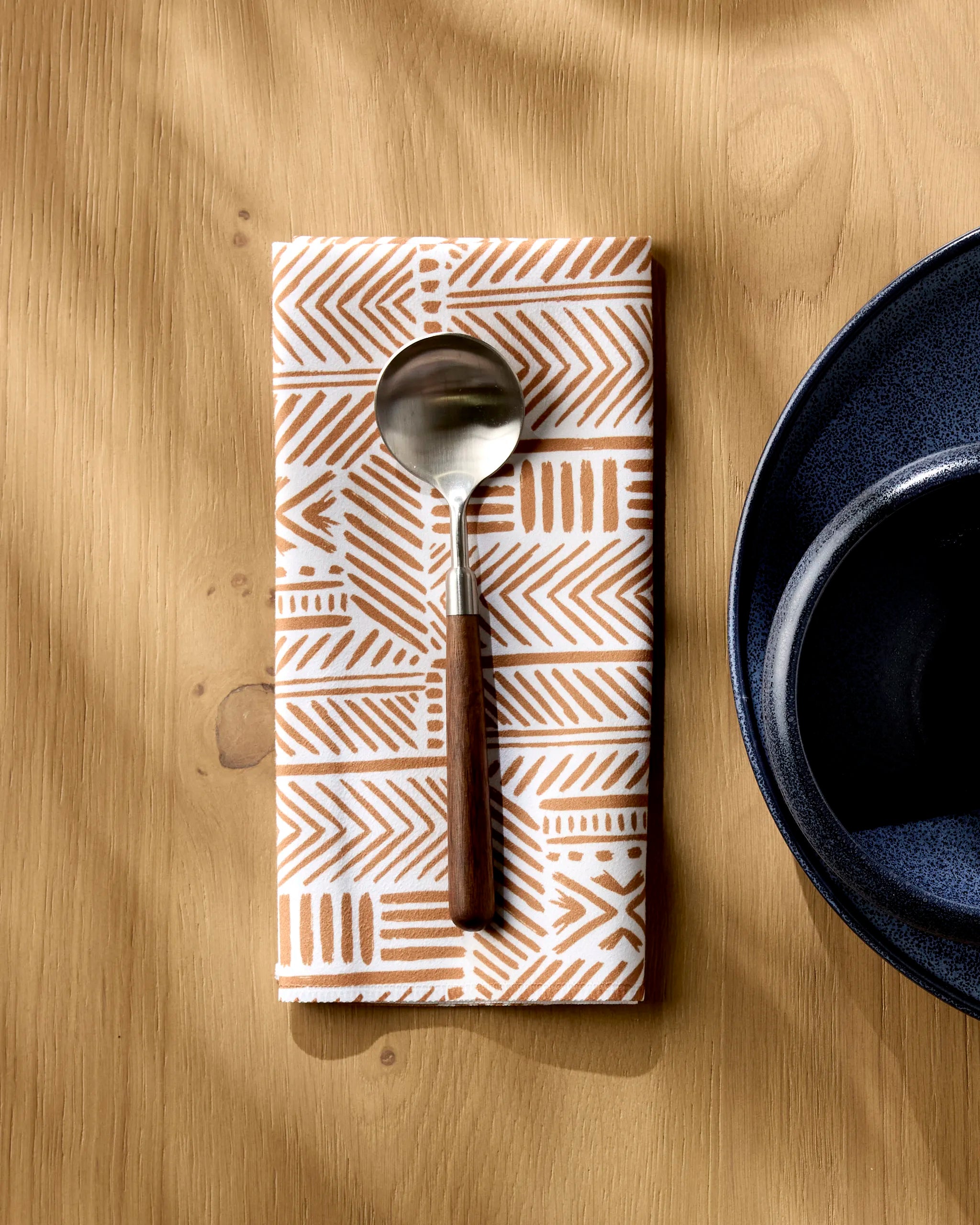 A spoon with a wooden handle rests on Hufton Studio's Mud Cloth Natural, an eco-friendly dinner napkin, atop a wooden table beside a dark blue bowl.
