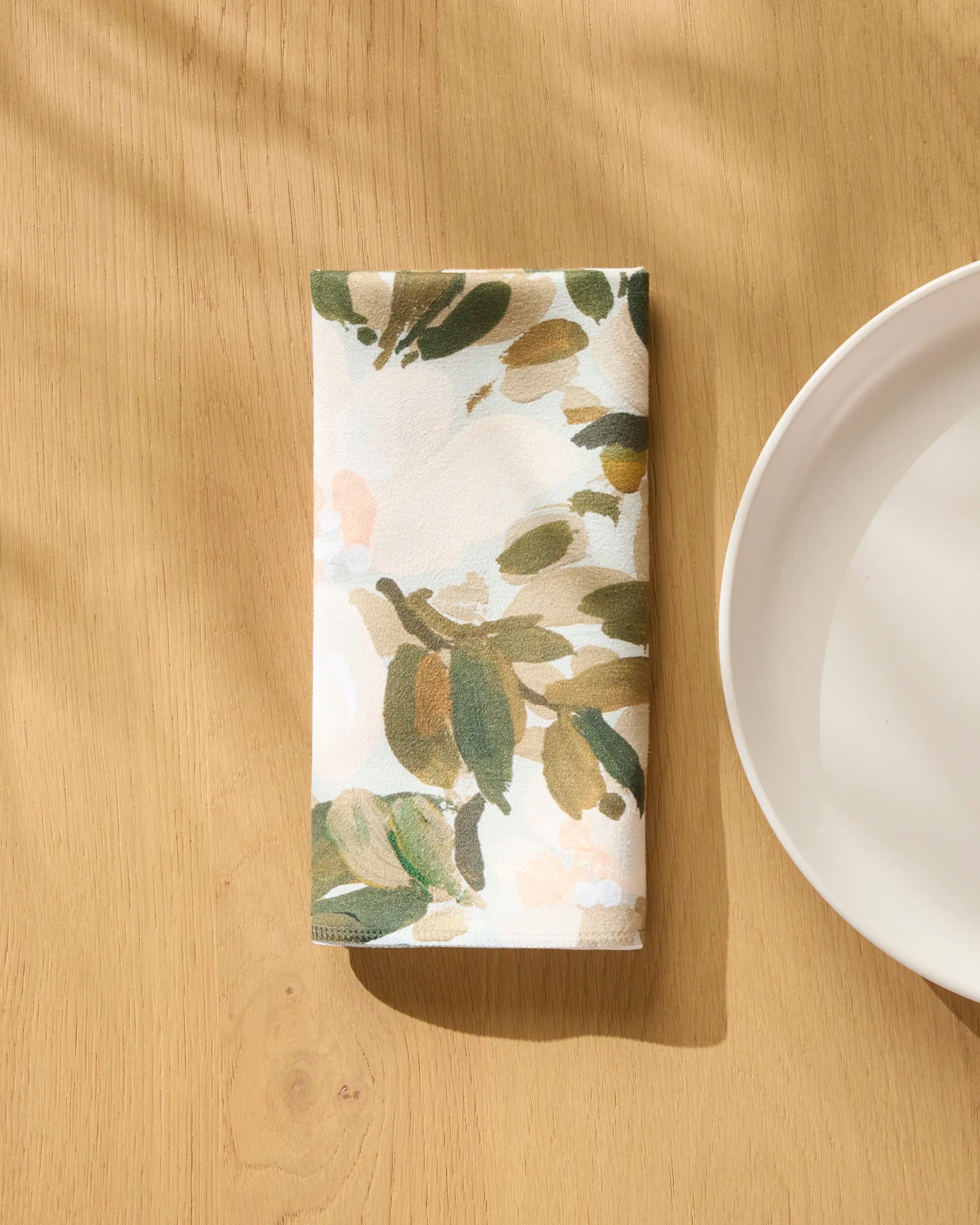 A Painted Blossoms reusable dinner napkin by Anee Shah, featuring a green and beige leafy design, lies folded on a light wooden table beside a white plate, with sunlight highlighting this elegant, eco-friendly tableware.