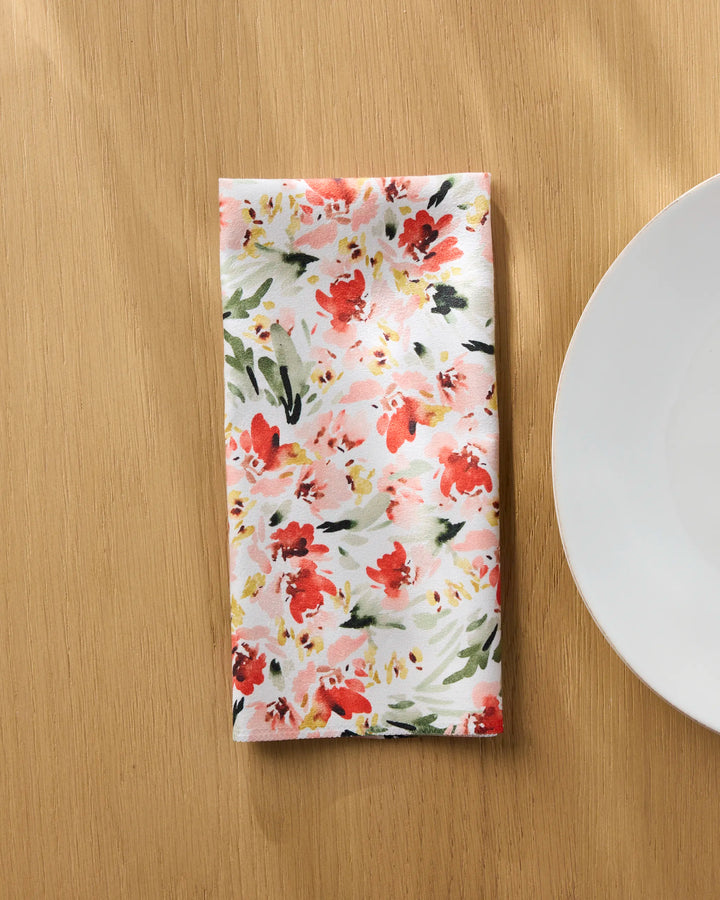The Melissa Johnson "Painted Garden" reusable napkin, featuring red and pink floral patterns on a light background, is neatly folded on a wooden table beside a white plate.