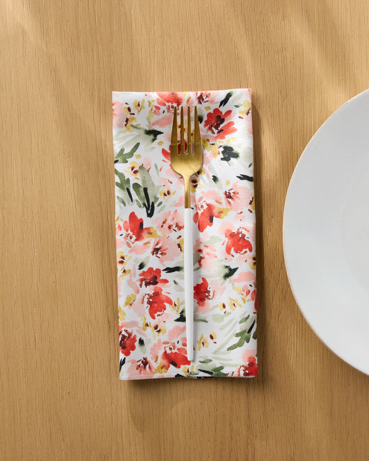 A gold fork rests on a folded Painted Garden reusable napkin by Melissa Johnson, featuring red, pink, and green floral patterns, set on a light wooden table next to a white plate.