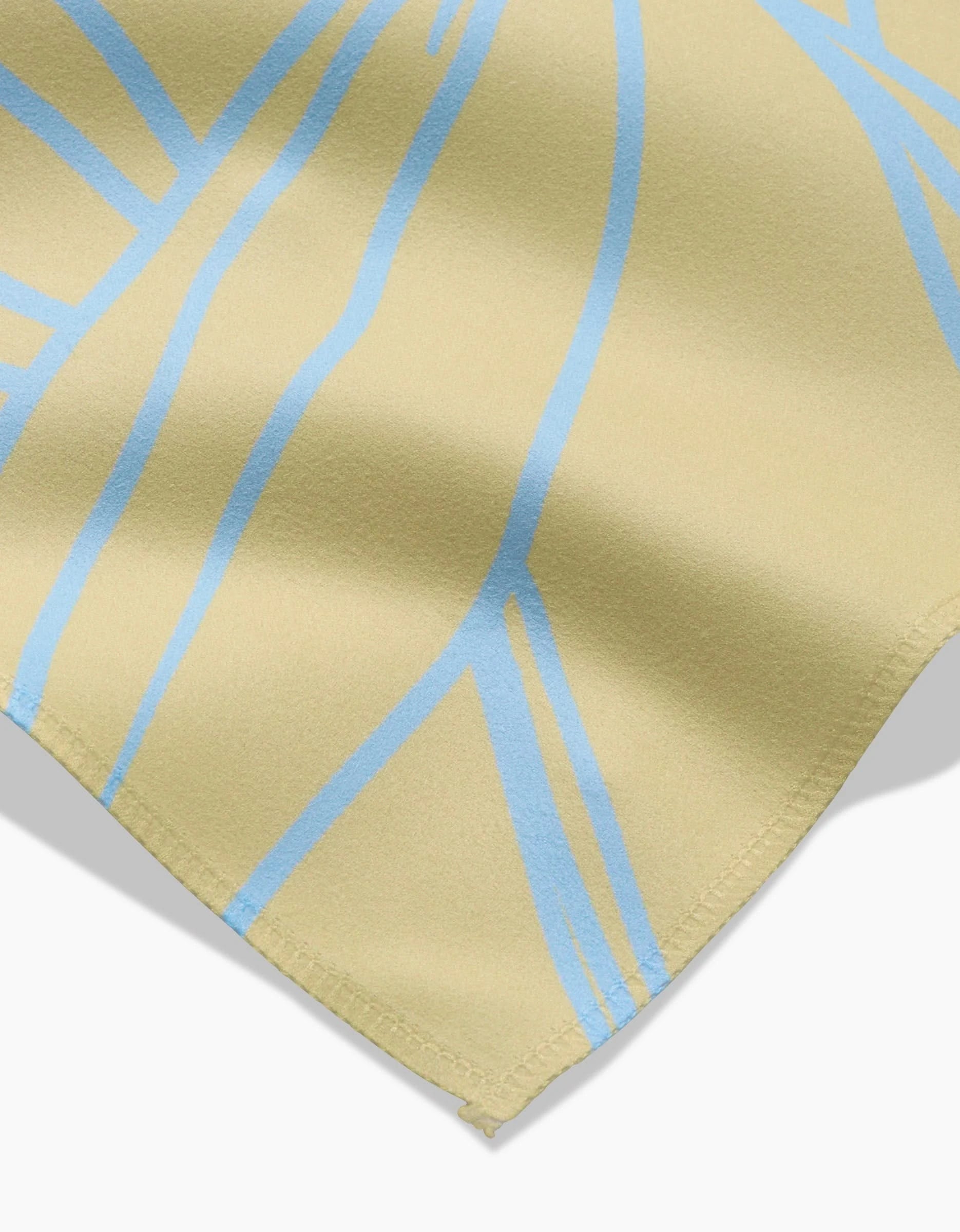 A close-up of the GEOMETRY Paradise Blooms dinner napkin in beige Geosuede fabric, featuring light blue abstract lines and a visible stitched edge, photographed on a white background.