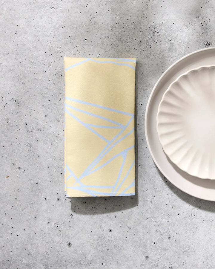 The GEOMETRY Paradise Blooms folded yellow washable napkin with light blue geometric lines rests on a gray speckled surface beside two stacked white scalloped-edge plates.