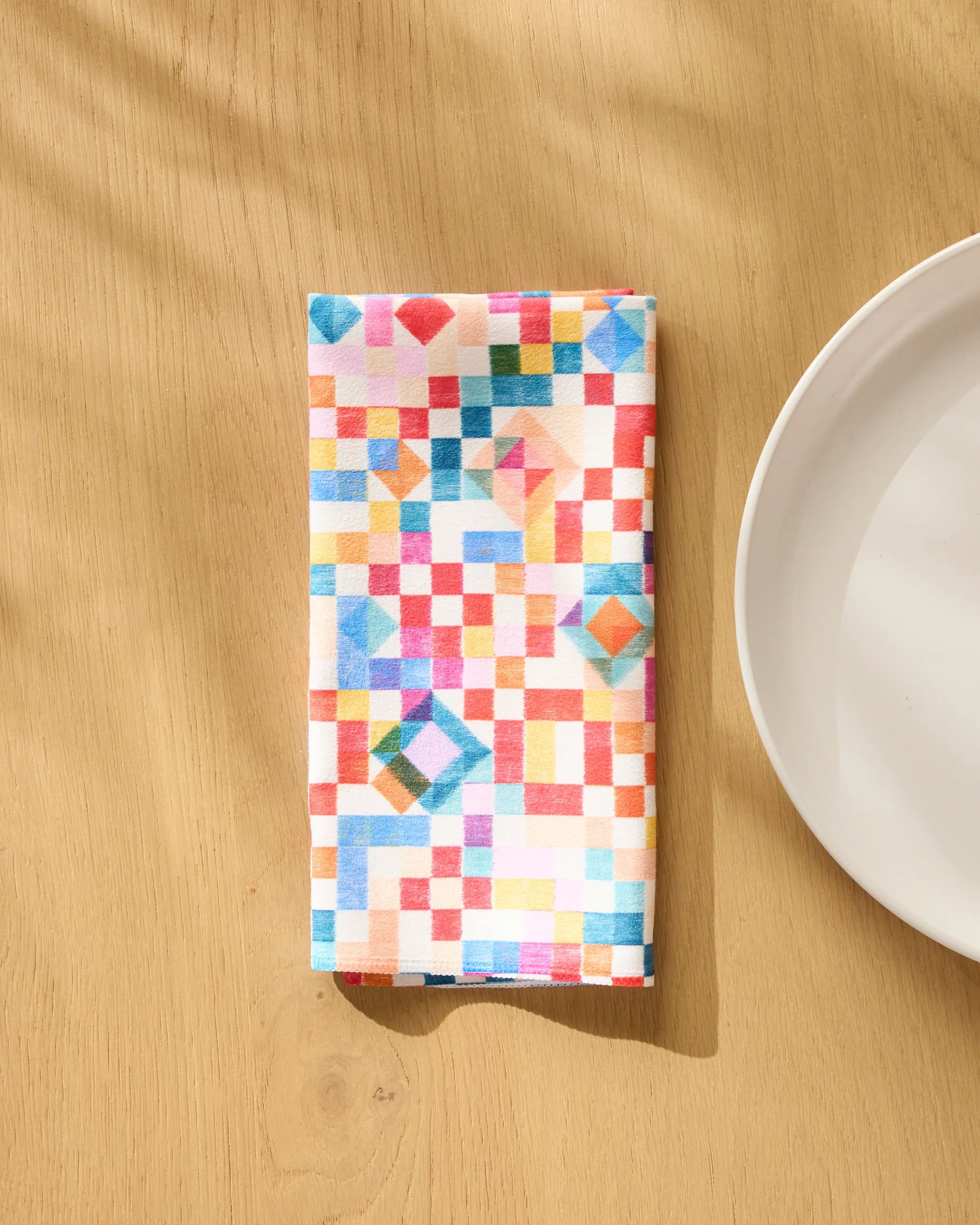 The Patchwork Geo reusable napkin by Rachel Parker features a vibrant geometric square pattern and sits on a light wooden table beside a white plate, adding charm to your Dinner Napkin Set.