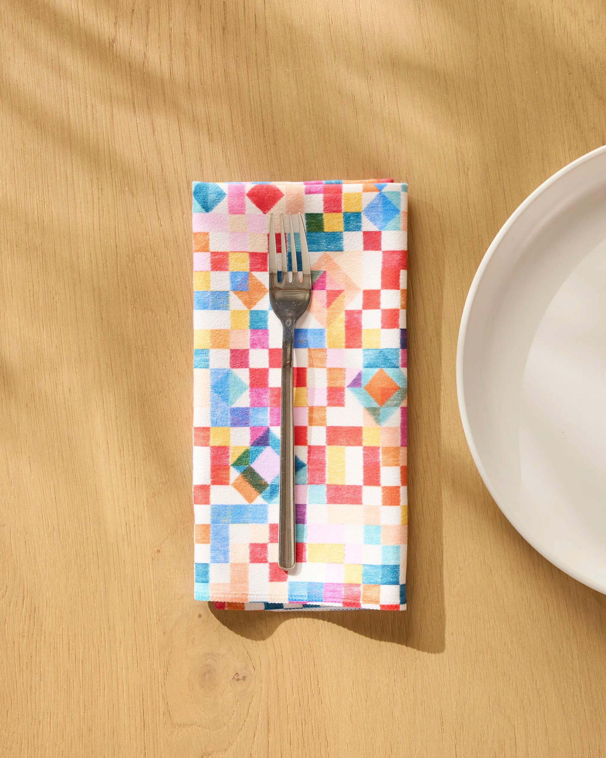 A silver fork rests on the Patchwork Geo dinner napkin set by Rachel Parker, featuring colorful geometric patterns, atop a light wooden table with part of a white plate visible to the right.