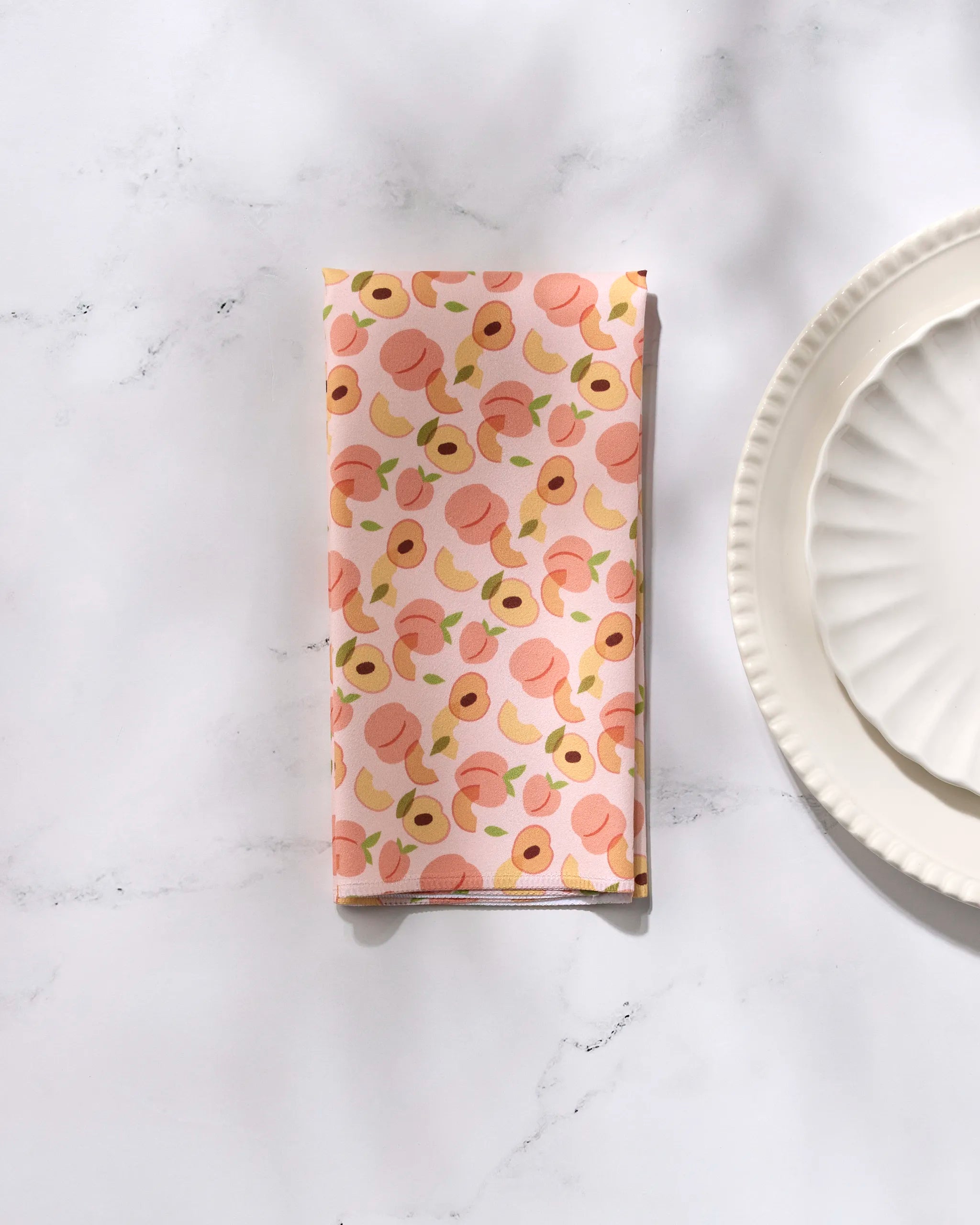 The GEOMETRY Peaches recycled dinner napkin, featuring a peach and leaf pattern, is folded on a marble surface beside two stacked white scalloped plates.