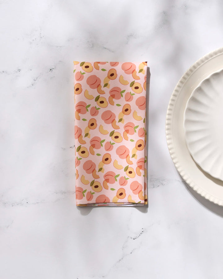 The GEOMETRY Peaches recycled dinner napkin, featuring a peach and leaf pattern, is folded on a marble surface beside two stacked white scalloped plates.