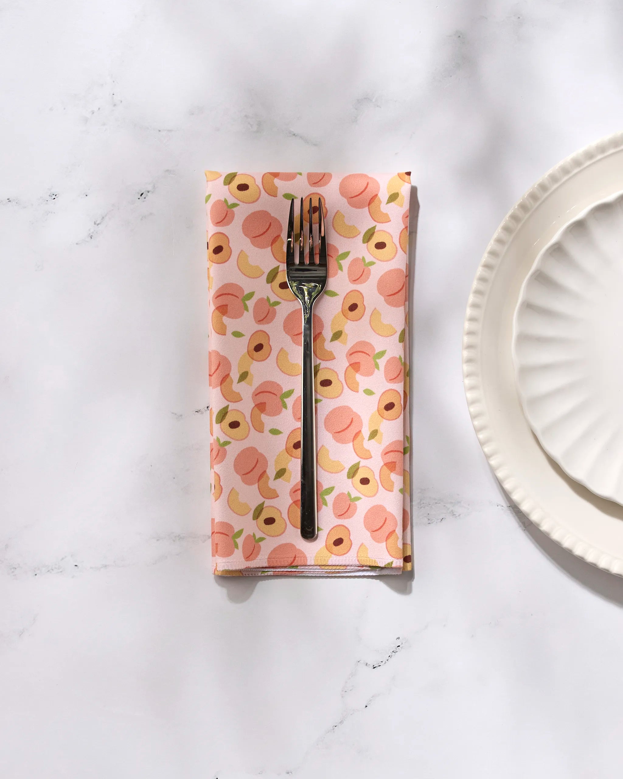 A silver fork rests on a folded GEOMETRY "Peaches" napkin, set beside white scalloped plates atop a white marble surface.