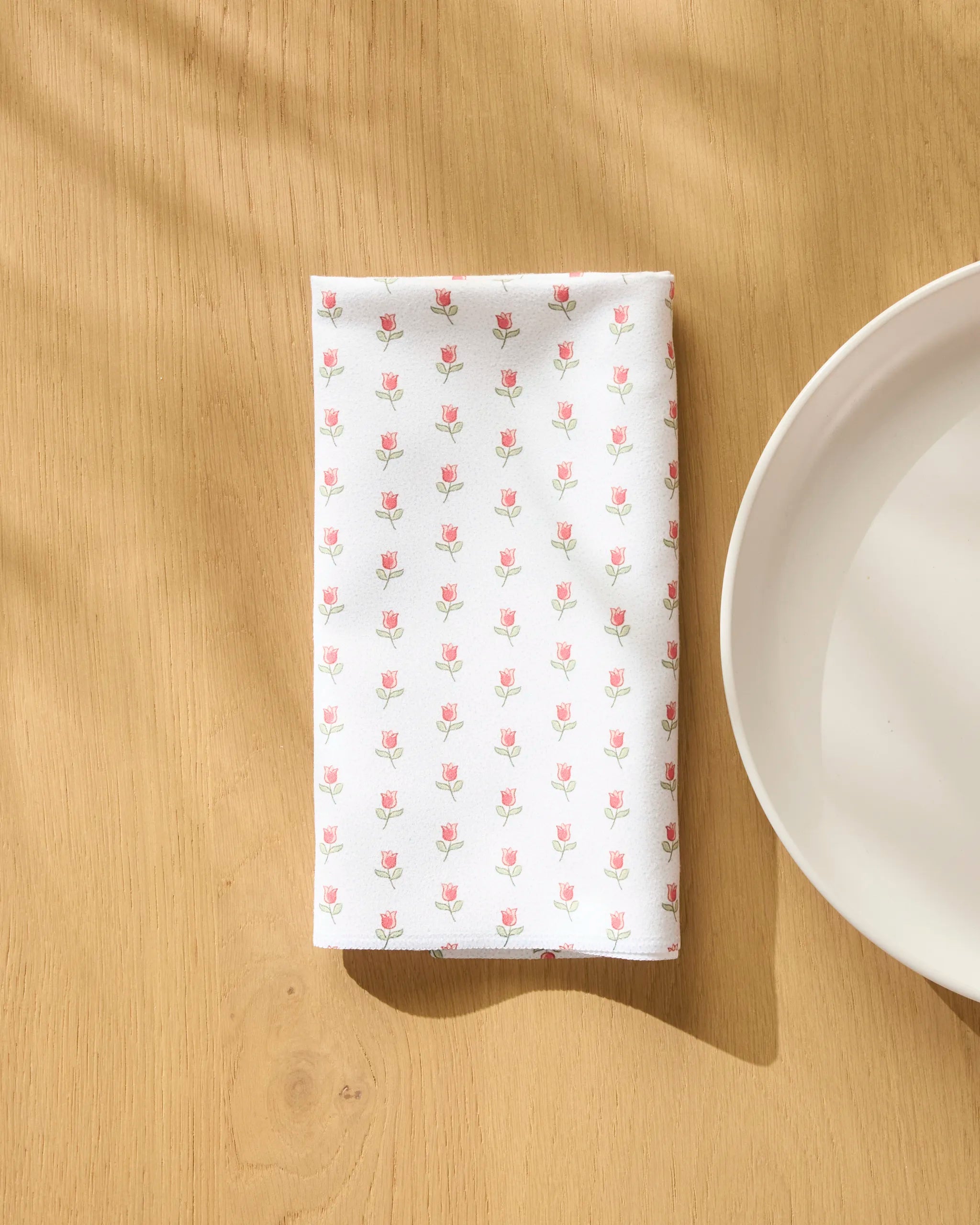 A Danelys Sidron Petite Tulip recycled dinner napkin with a small pink tulip pattern lies folded on light wood beside a white plate, soft sunlight casting gentle shadows across the table.