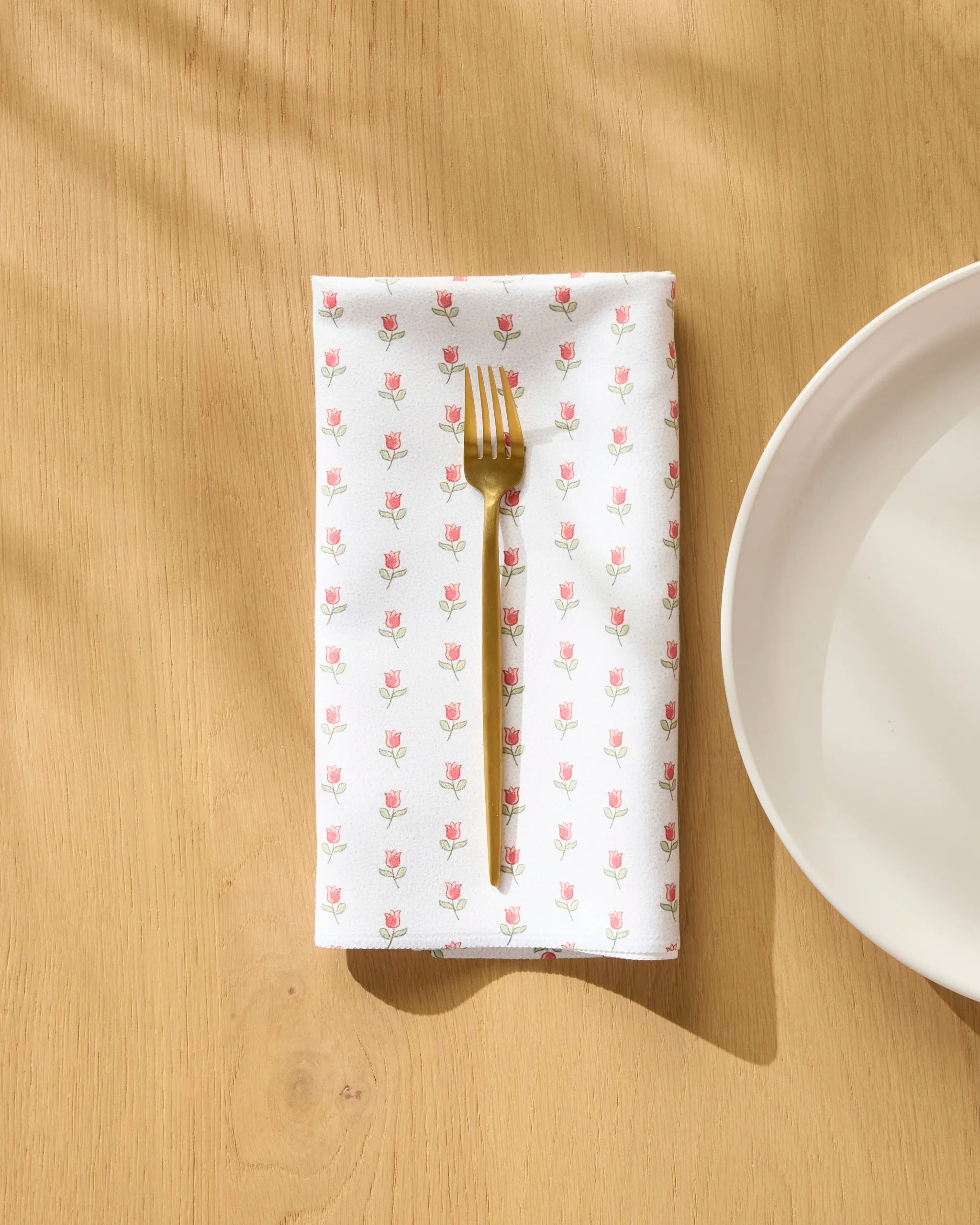 A gold fork rests on the Danelys Sidron Petite Tulip white reusable napkin, featuring a pink tulip pattern, set on a light wooden table beside a white plate.