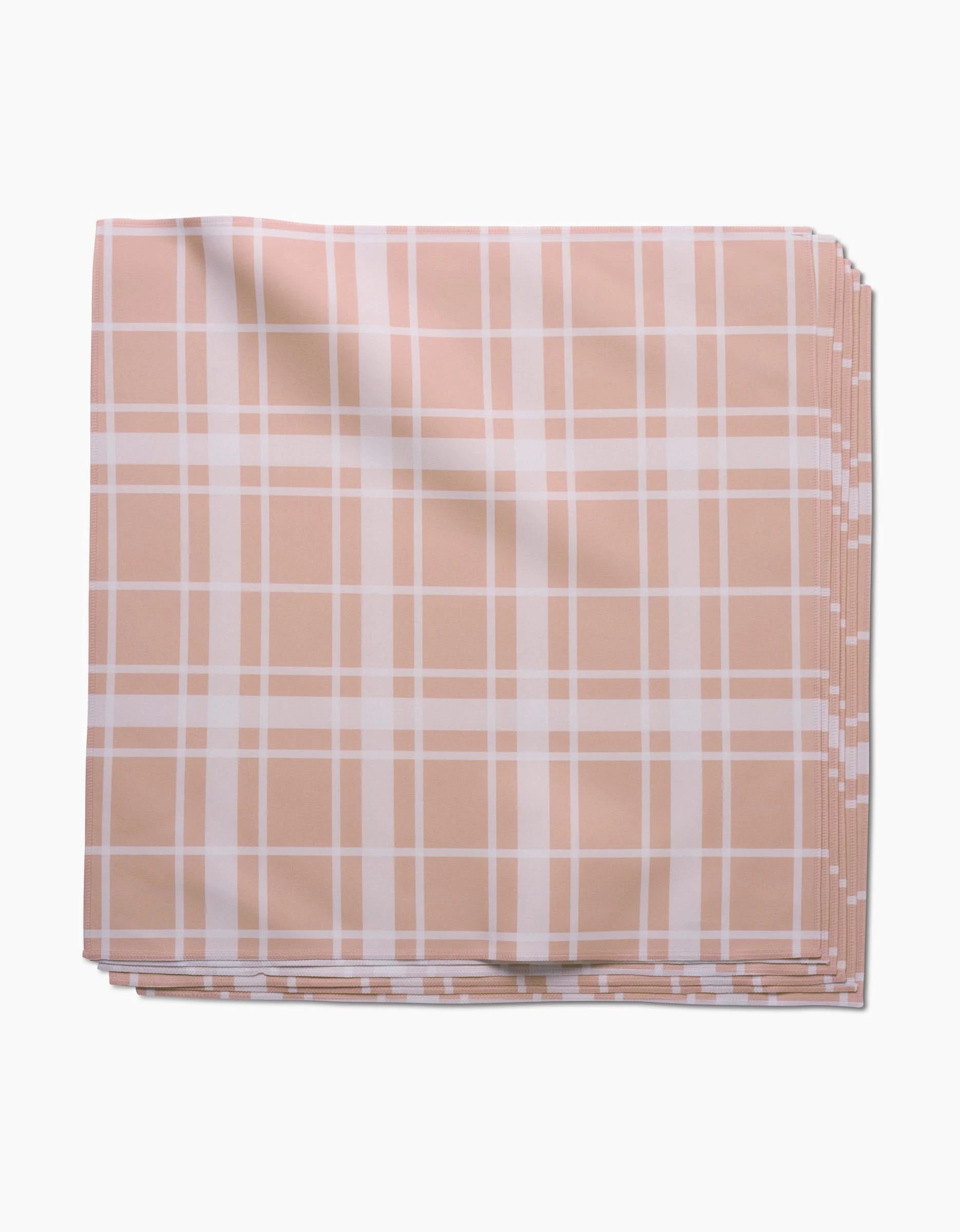 Aubrey Fairchild’s Picnic Time Pink dinner napkin, neatly folded, features soft Geosuede fabric with a peach background and white plaid pattern, shown on a white surface.