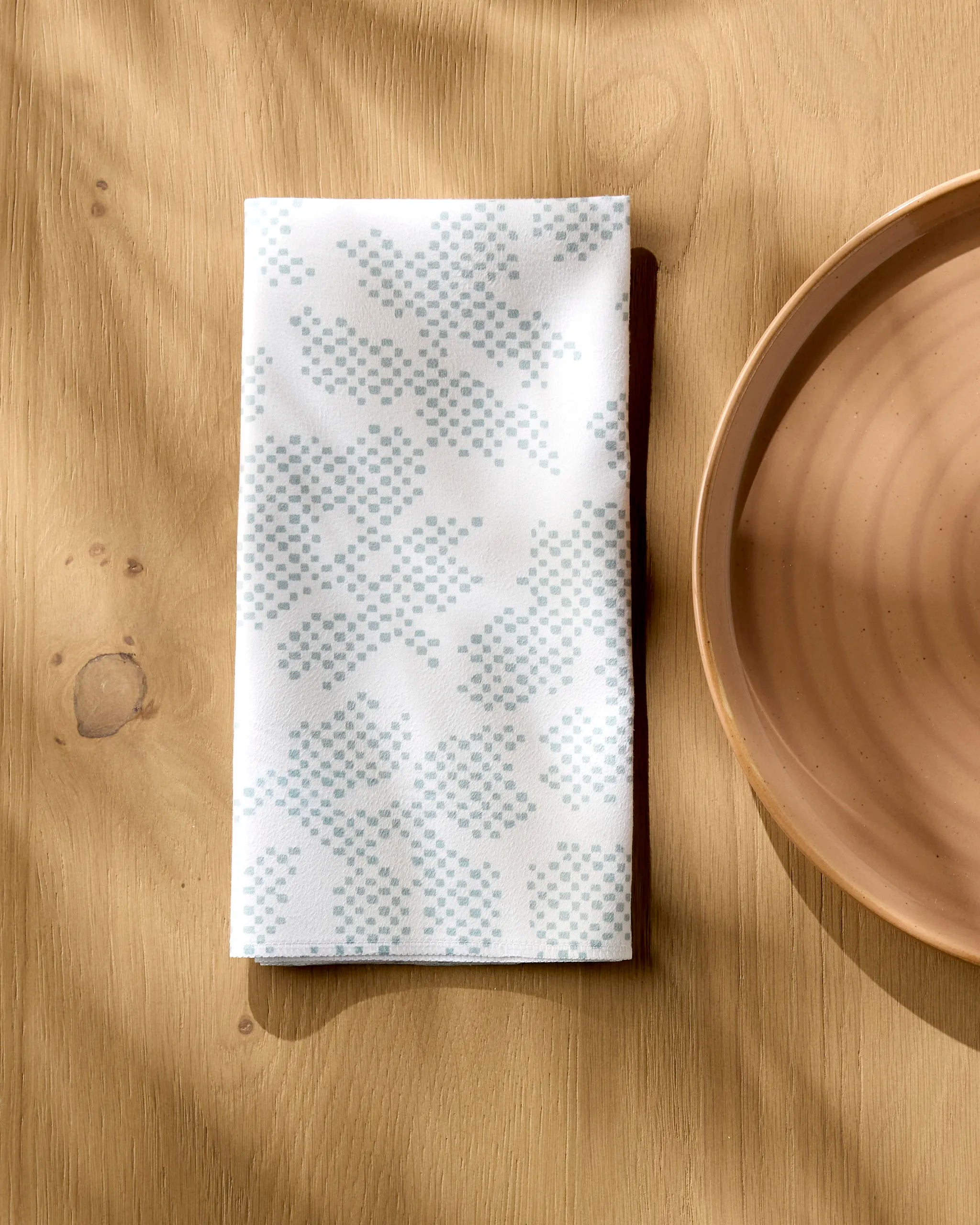 The Pixie Sage reusable white cloth napkin by Studio Jeannot, featuring a light blue dotted pattern, rests on a wooden table beside a light brown ceramic plate as sunlight casts gentle shadows.