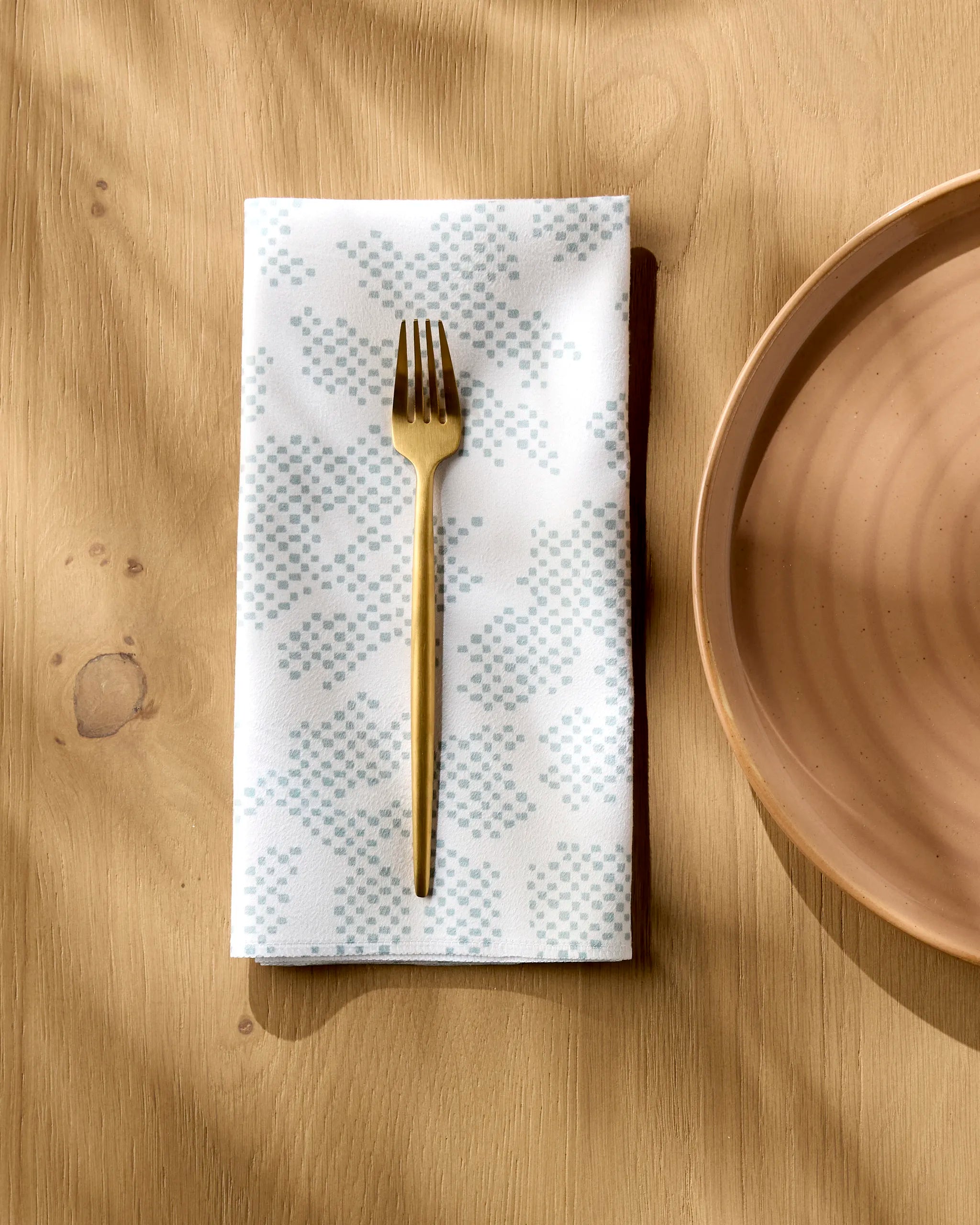 A gold fork sits on Studio Jeannot’s Pixie Sage reusable cloth napkin with a light blue pattern, next to a light brown ceramic plate on a wooden table.