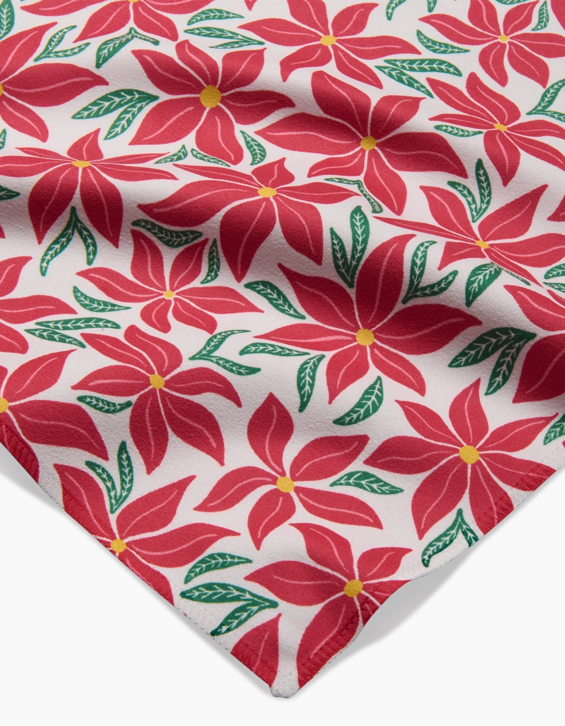 A close-up of Kayla Ann’s Poinsettia Party napkin, featuring red poinsettias, green leaves, and yellow centers on white with gentle folds—part of our recycled Dinner Napkin Set.