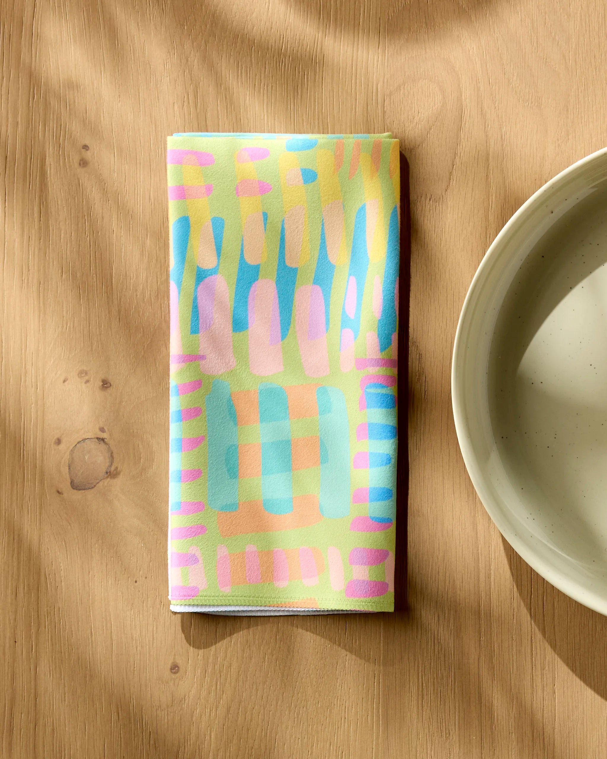 The "Positive Vibes" reusable napkin by Sareka Unique, featuring a colorful abstract blue, yellow, and pink design, is folded on a wooden table beside a white ceramic plate.