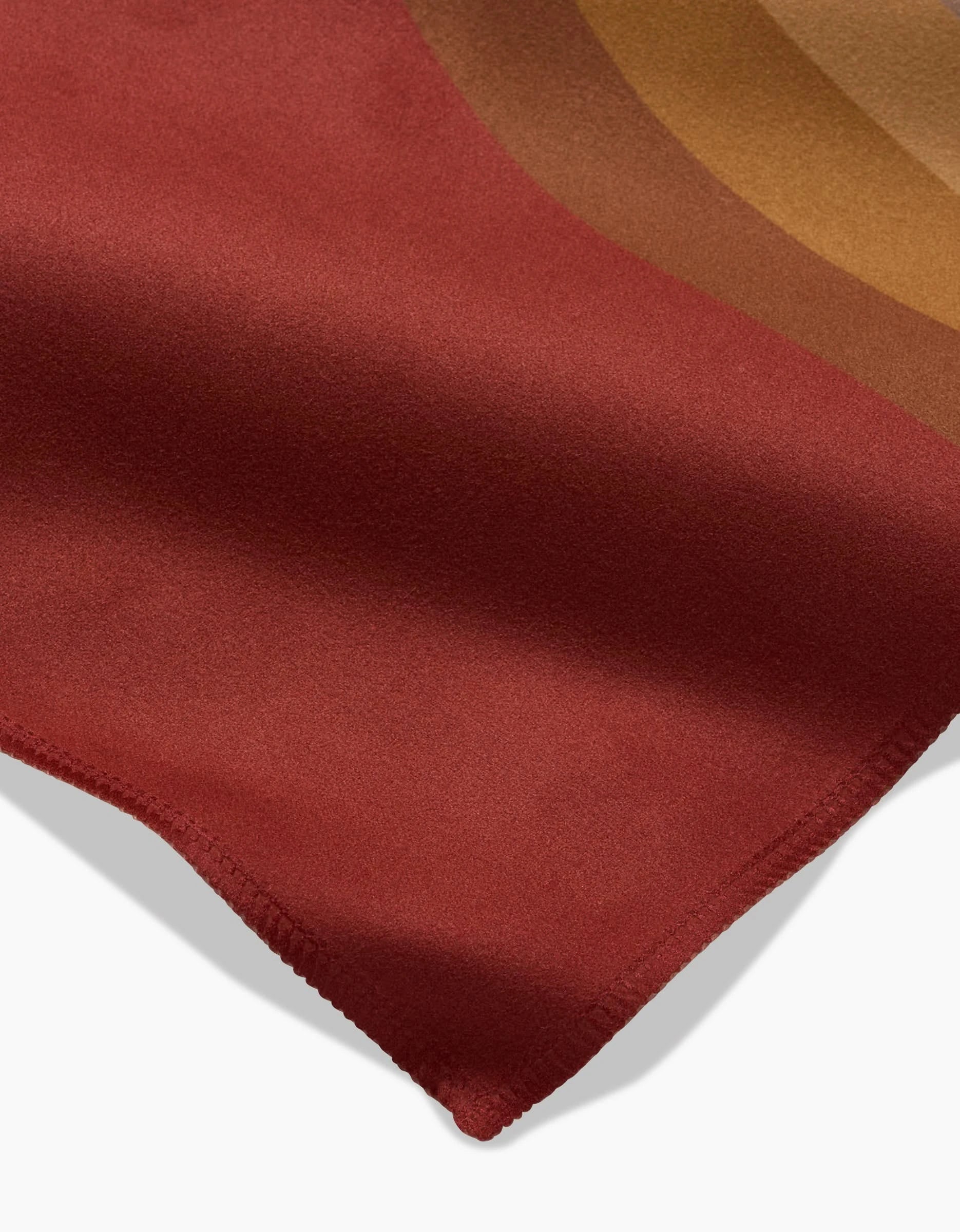 A close-up of the GEOMETRY Pumpkin Spice napkin, featuring soft red fabric with curved brown and gold bands in the corner and a neat zigzag-stitched edge, set against a white background.