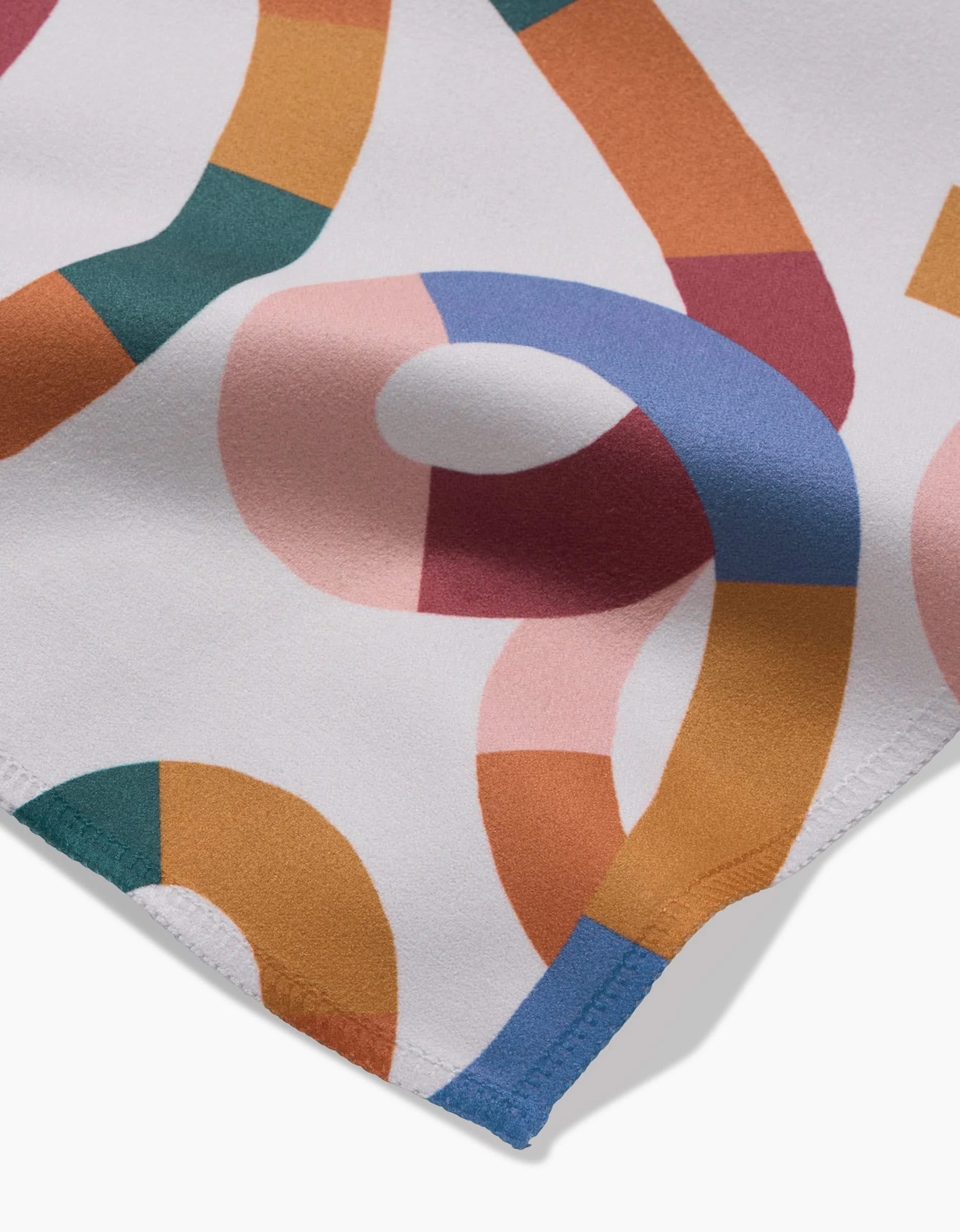 A close-up of Claire Iglesias’ Rainbow Squiggles dinner napkins shows white Geosuede fabric with colorful, abstract intertwining bands and visible hem stitching. These eco-friendly napkins feature orange, pink, green, blue, and mustard hues.