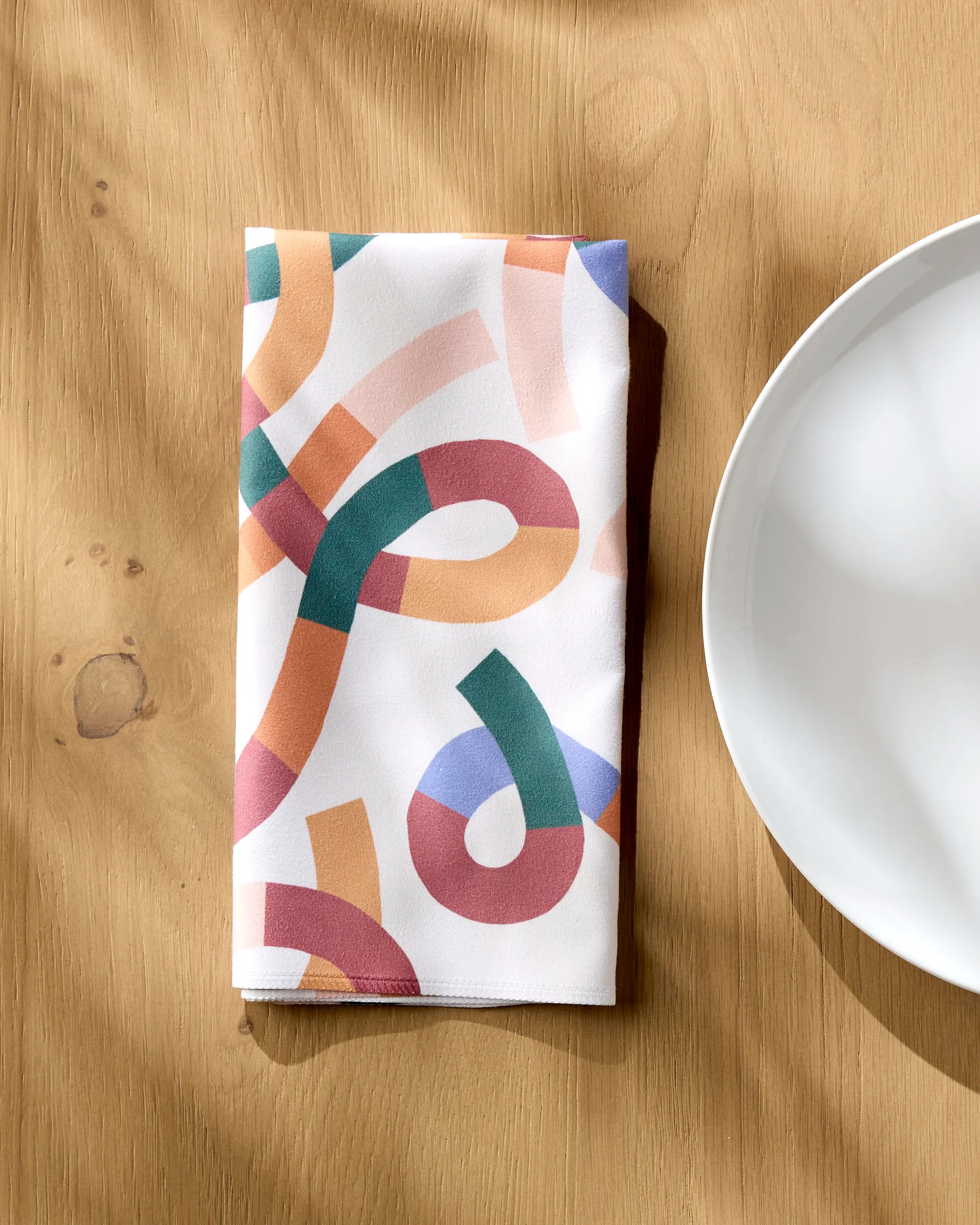 The Rainbow Squiggles reusable napkin by Claire Iglesias, featuring a colorful abstract pattern, is folded on a wooden table beside a white plate, with soft shadows cast across the scene.