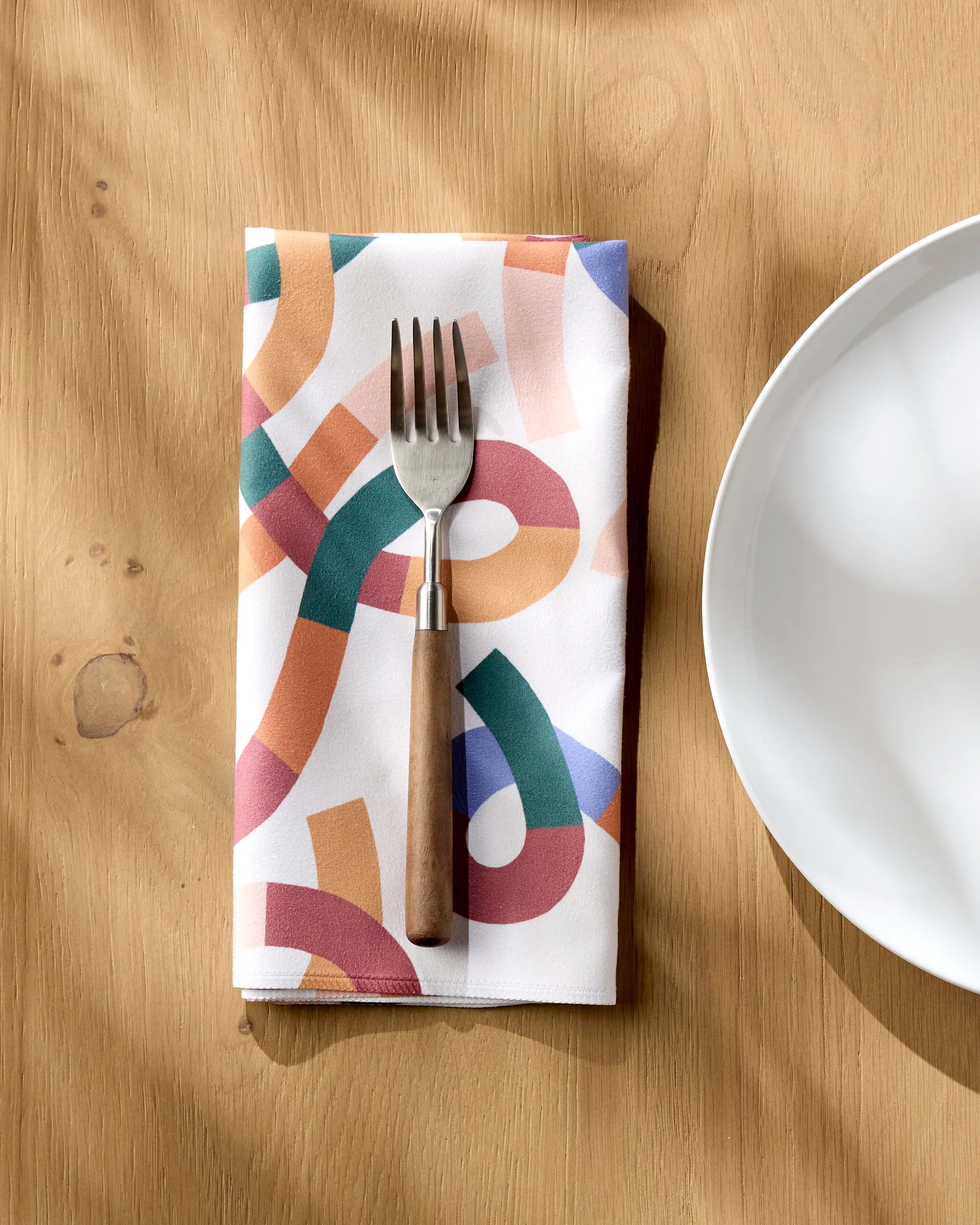 The Claire Iglesias Rainbow Squiggles reusable napkin lies beside a white plate on a light wooden table, with a metal fork featuring a wooden handle resting on it as sunlight casts soft shadows across the scene.