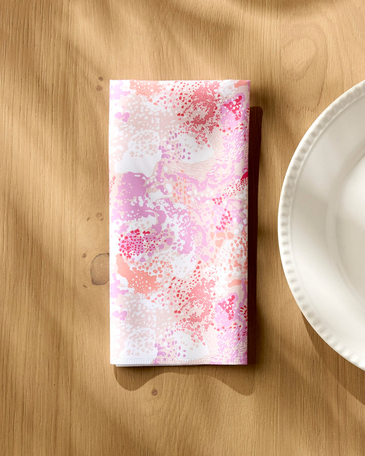 A folded white "Rawhide" reusable dinner napkin by Elisabeth Wing, featuring a pink and red abstract floral pattern, rests on a wooden table beside a white plate, illuminated by sunlight and soft shadows.