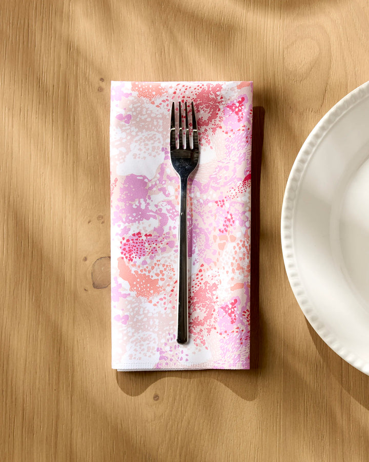 A fork rests on a Rawhide reusable dinner napkin by Elisabeth Wing, featuring a pink and white pattern, neatly folded on a wooden table beside the edge of a white plate.