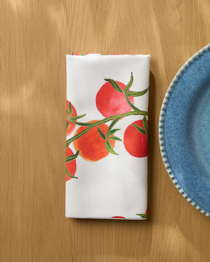 The Geometry "Ripe and Ready" folded white reusable table napkin with tomato print rests on a light wooden table beside a blue textured-rim ceramic plate.