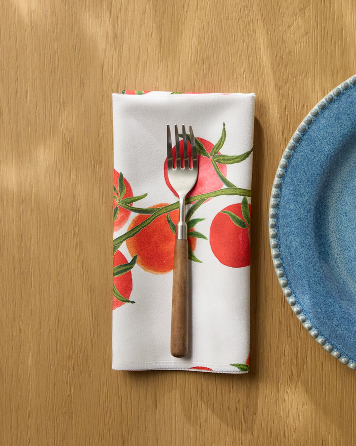 A fork with a wooden handle rests on Geometry's "Ripe and Ready" reusable table napkin, featuring red tomatoes and green leaves, placed on a light wooden table next to a blue ceramic plate.